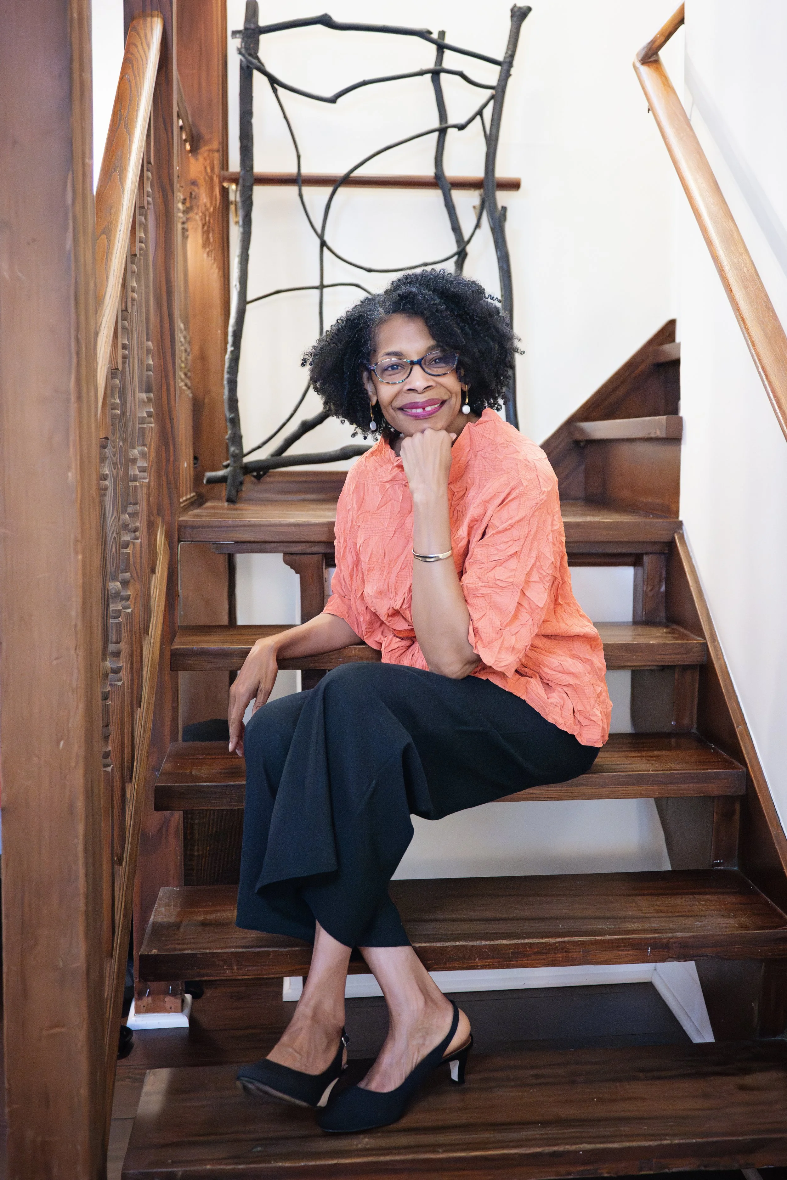 A woman with curly black hair, glasses, and earrings sitting on wooden stairs, smiling and resting her chin on her hand, wearing an orange blouse, black pants, and black heels.