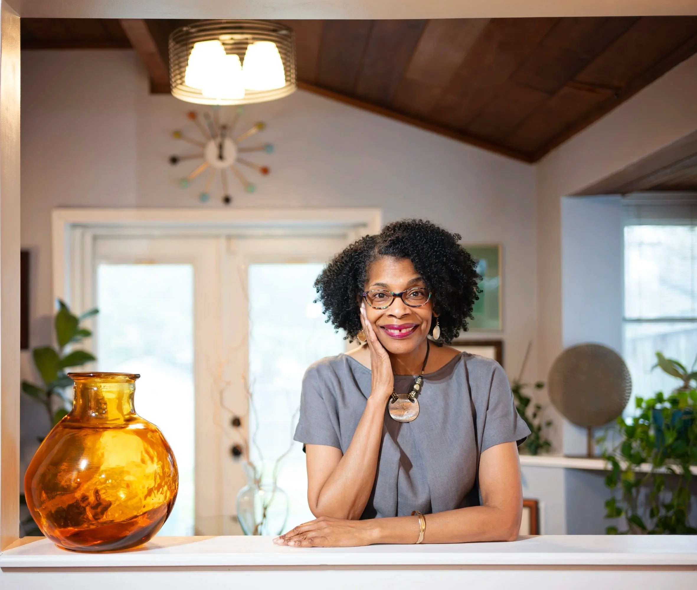 A woman with curly hair and glasses smiling, with her hand resting on her face, sitting at a table. There is an orange-glazed vase on the table and plant leaves in the foreground. The background features large windows, a ceiling light, and a modern wall clock.