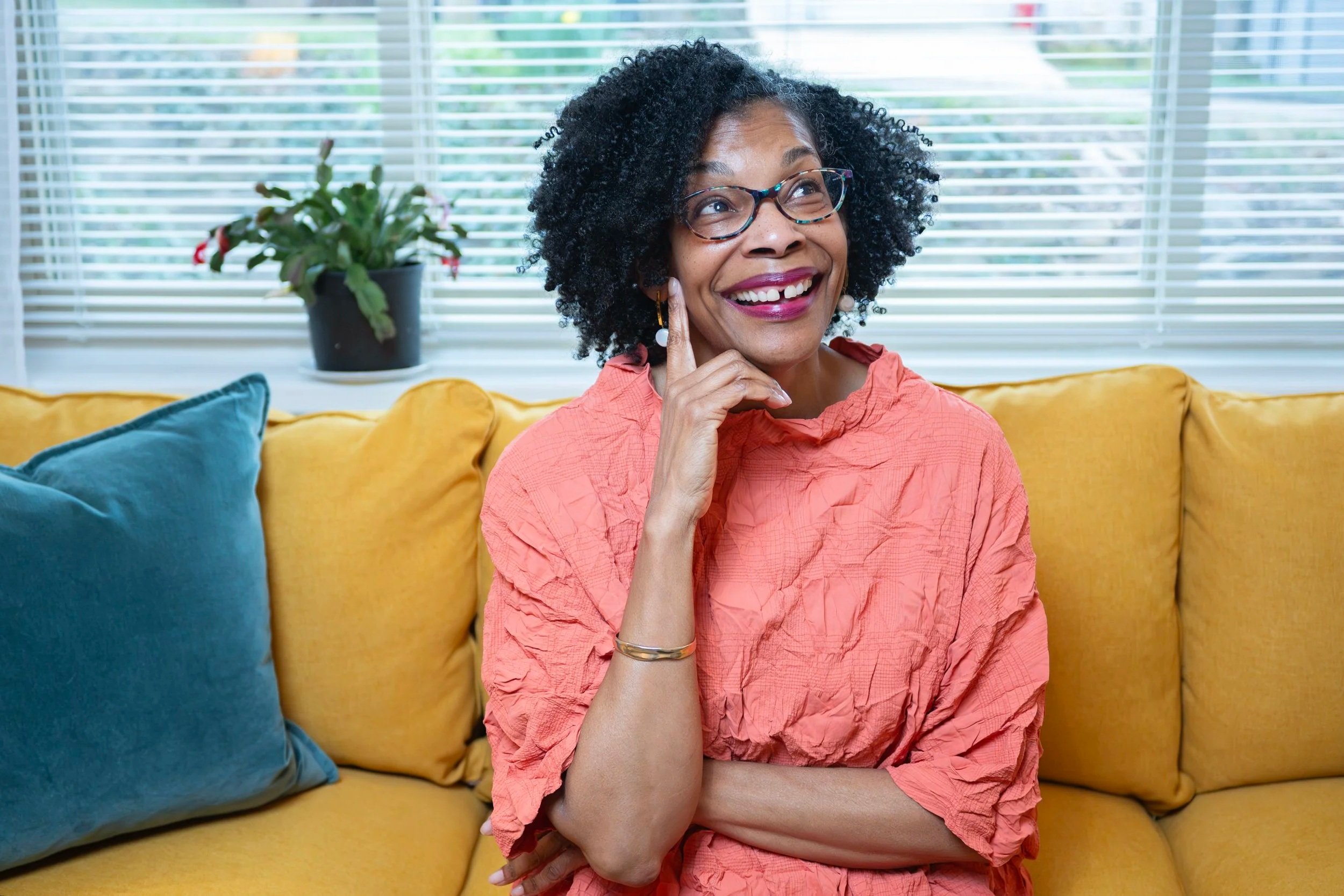 A woman with glasses and curly hair smiling, touching her chin, sitting on a yellow couch with teal and yellow pillows, in front of a window with blinds and a potted plant in the background.
