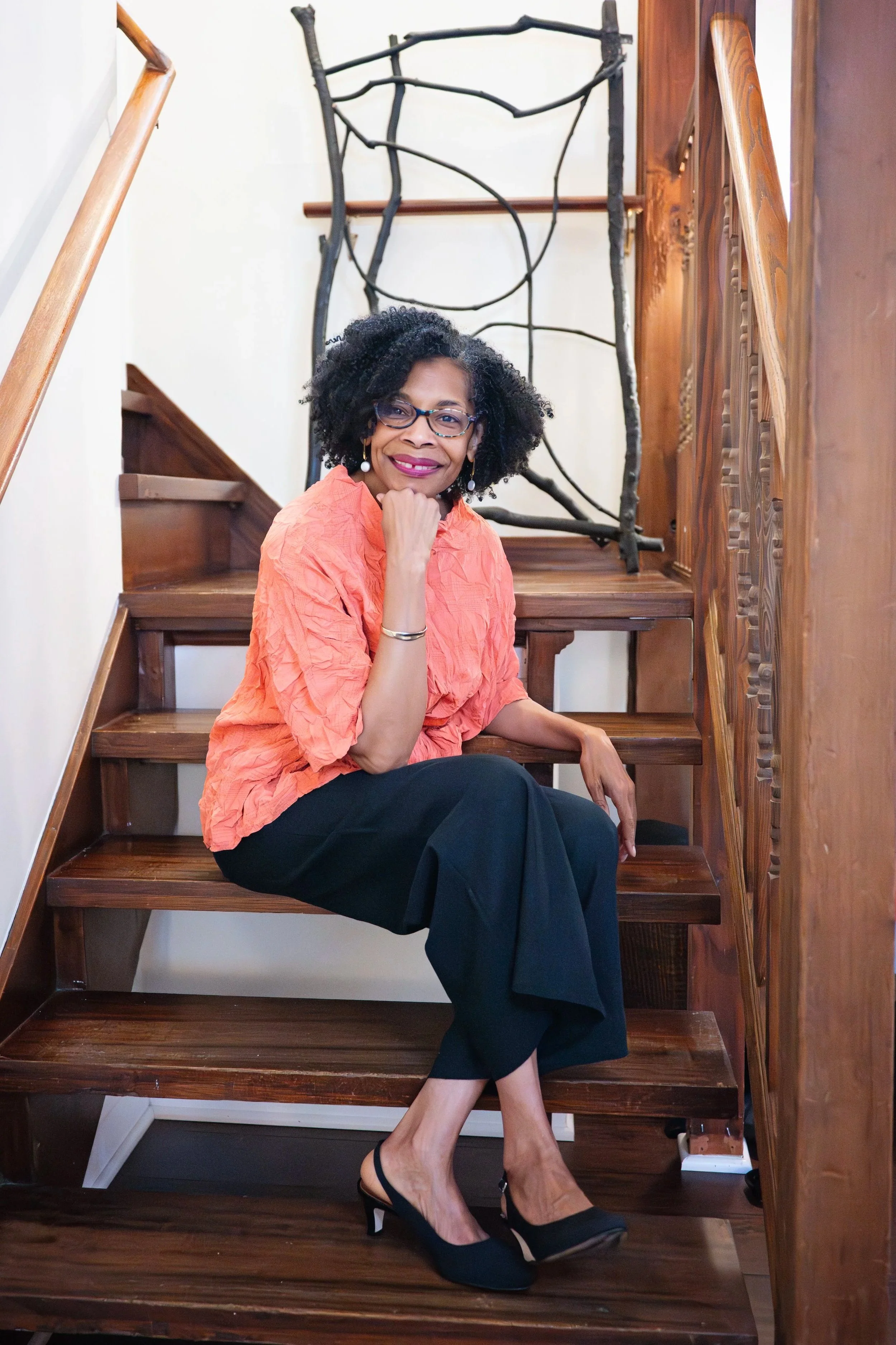 Woman sitting on wooden staircase with dark hair and glasses, wearing an orange blouse and black pants, smiling and resting her chin on her hand.