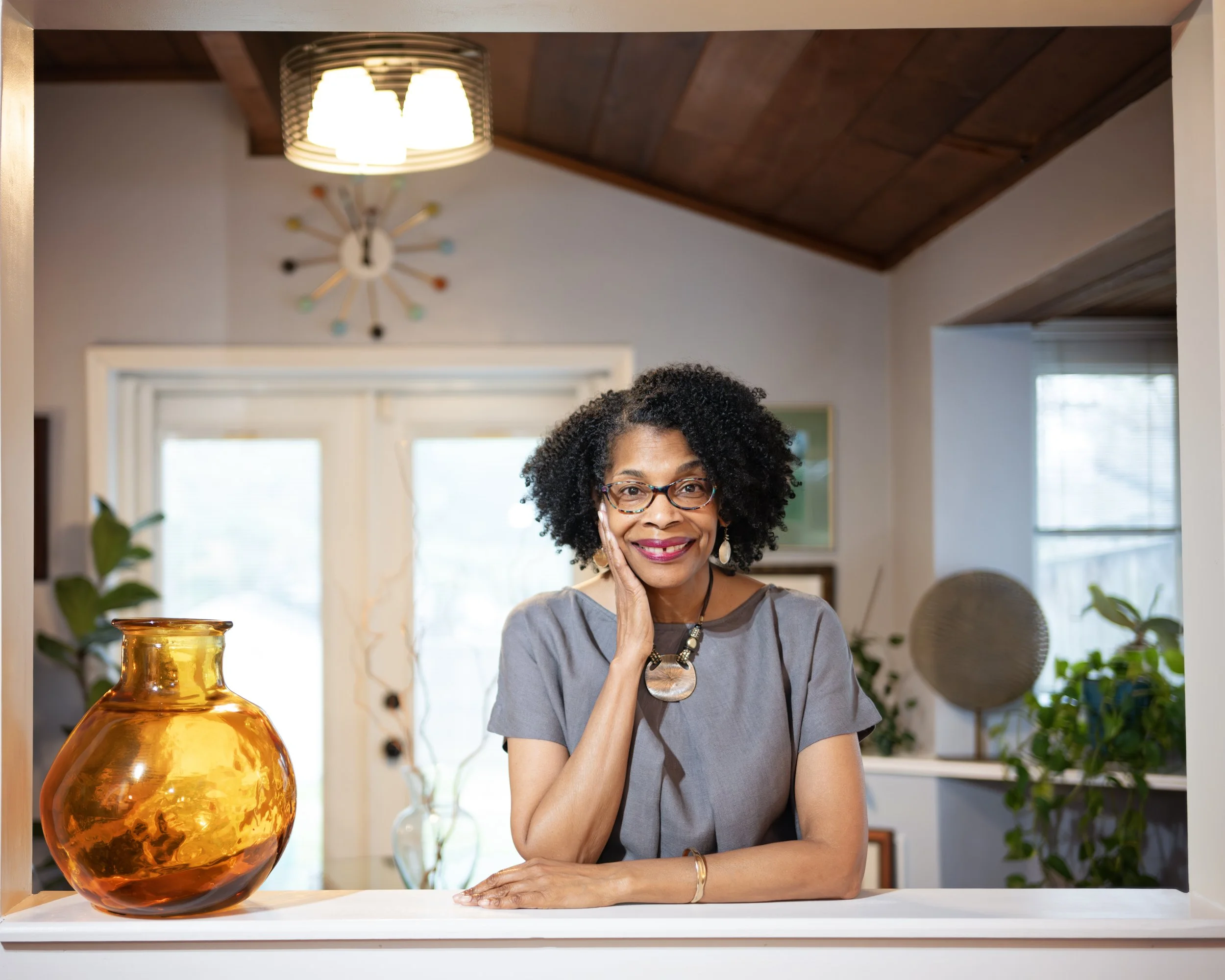 A middle-aged woman with glasses and curly hair smiling while sitting at a white table in a well-lit room with a large amber glass vase, potted plants, and a window in the background.