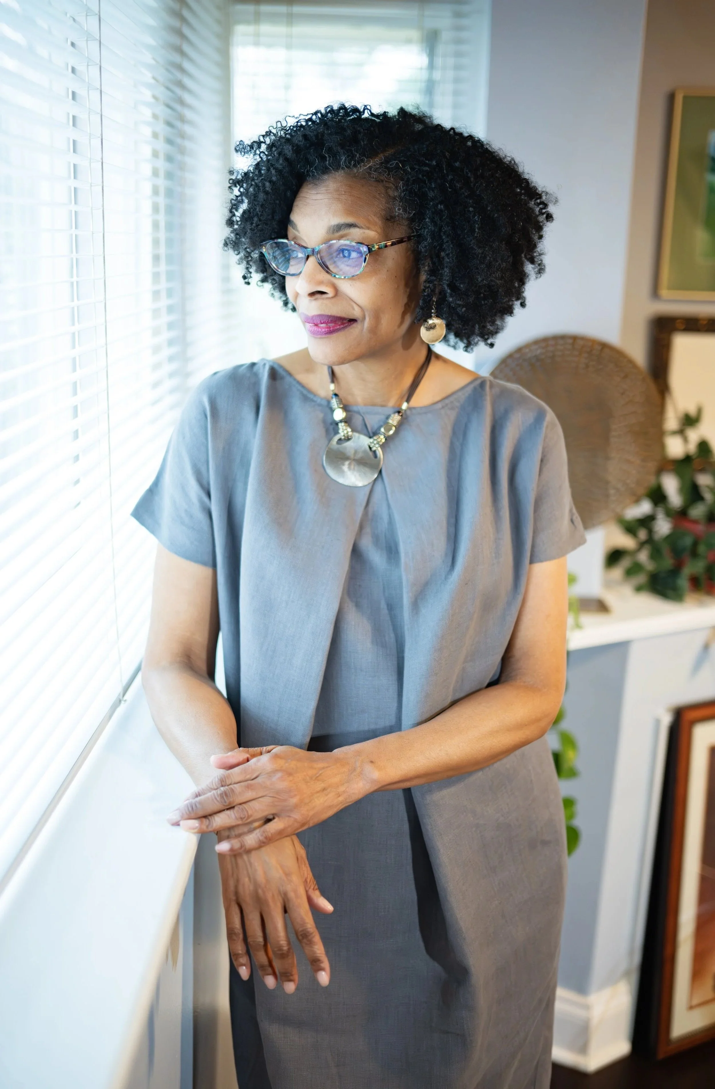 An older woman with curly black hair, glasses, and large earrings, standing indoors near a window with blinds, looking out thoughtfully.