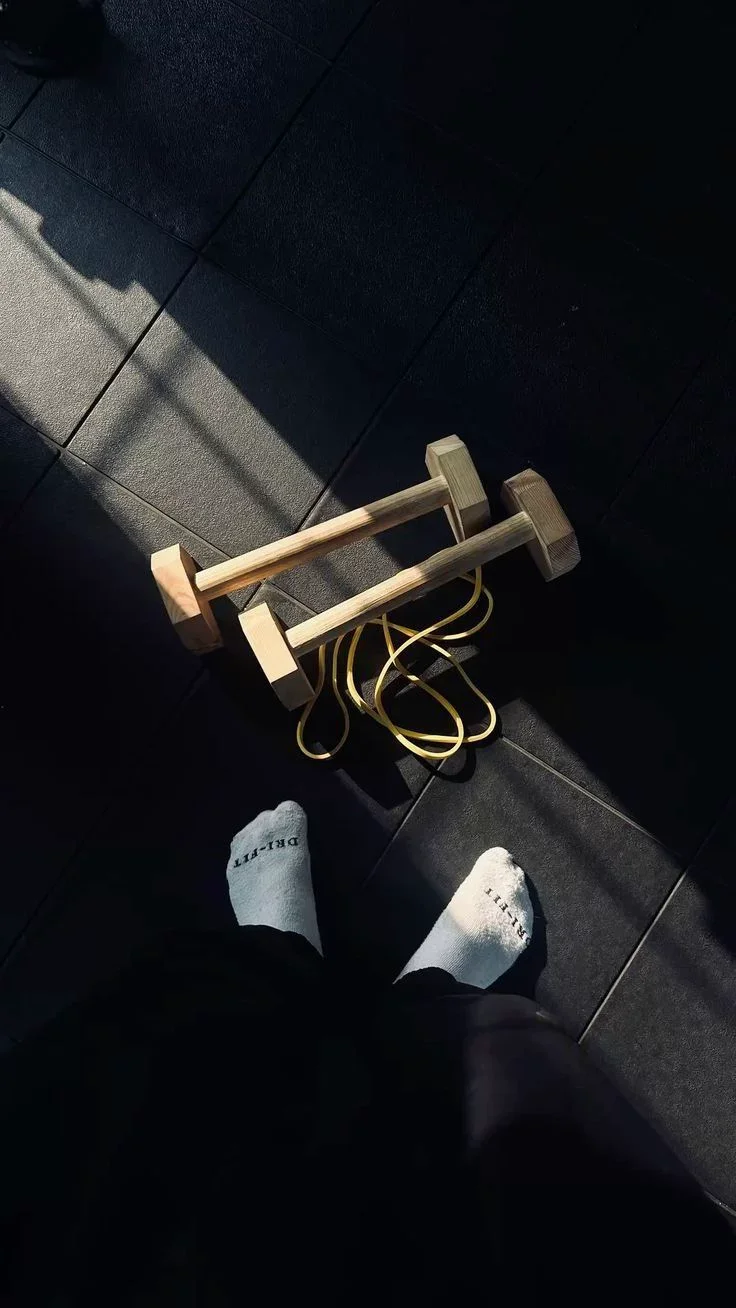A pair of feet wearing white socks with black pants, standing on a dark tiled floor. Near the feet is a small wooden stool with yellow elastic bands draped over it, and a shadow cast on the floor.