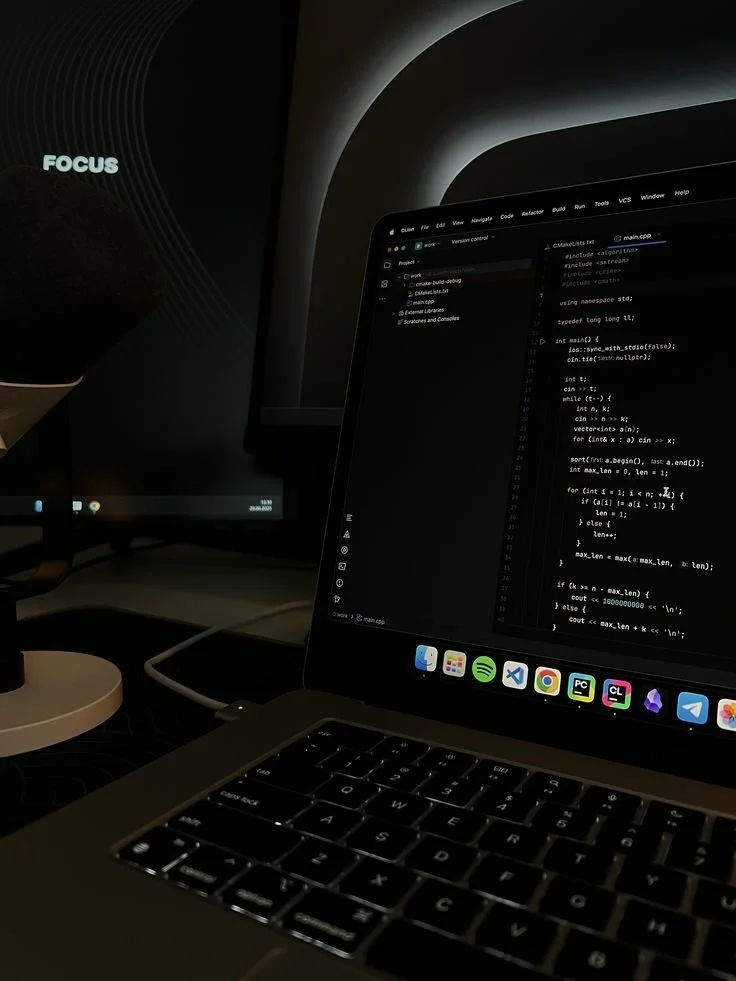 A laptop displaying a C++ programming code in a dark room with a monitor showing the word 'FOCUS' on the left side.