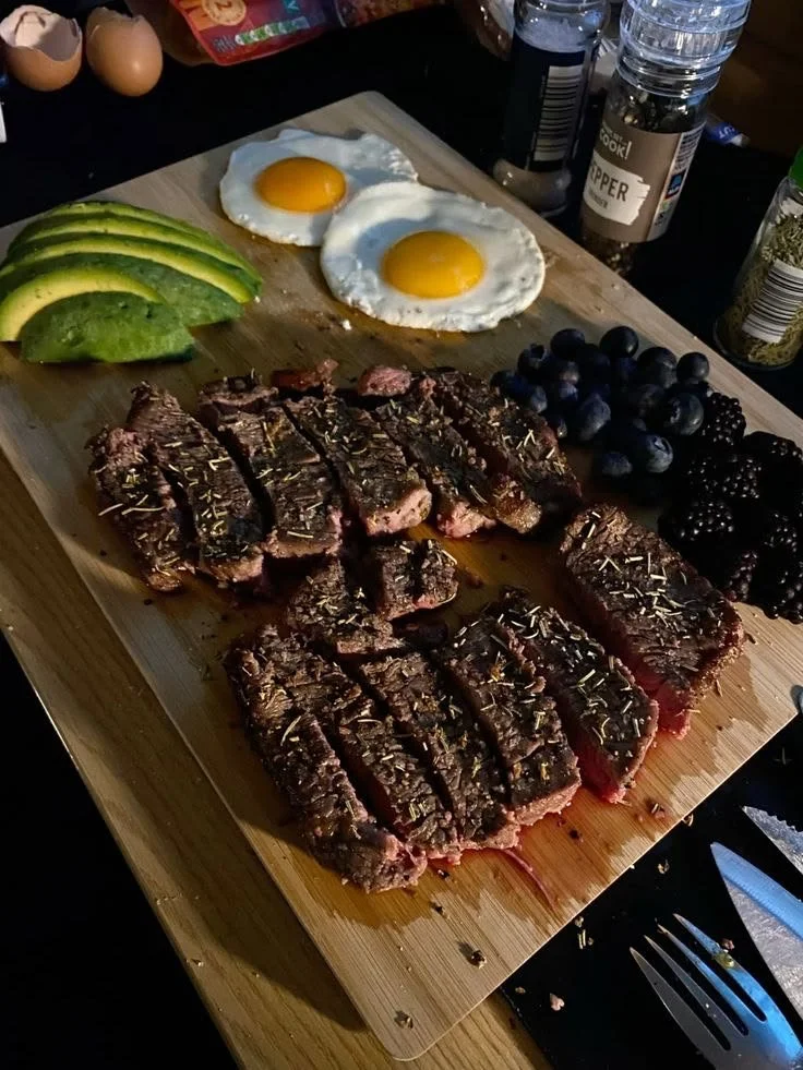 Steak cooked to medium rare topped with herbs, sliced and served on a wooden cutting board with avocado slices, two fried eggs, and a variety of blackberries and blueberries, with salt and pepper shakers in the background.