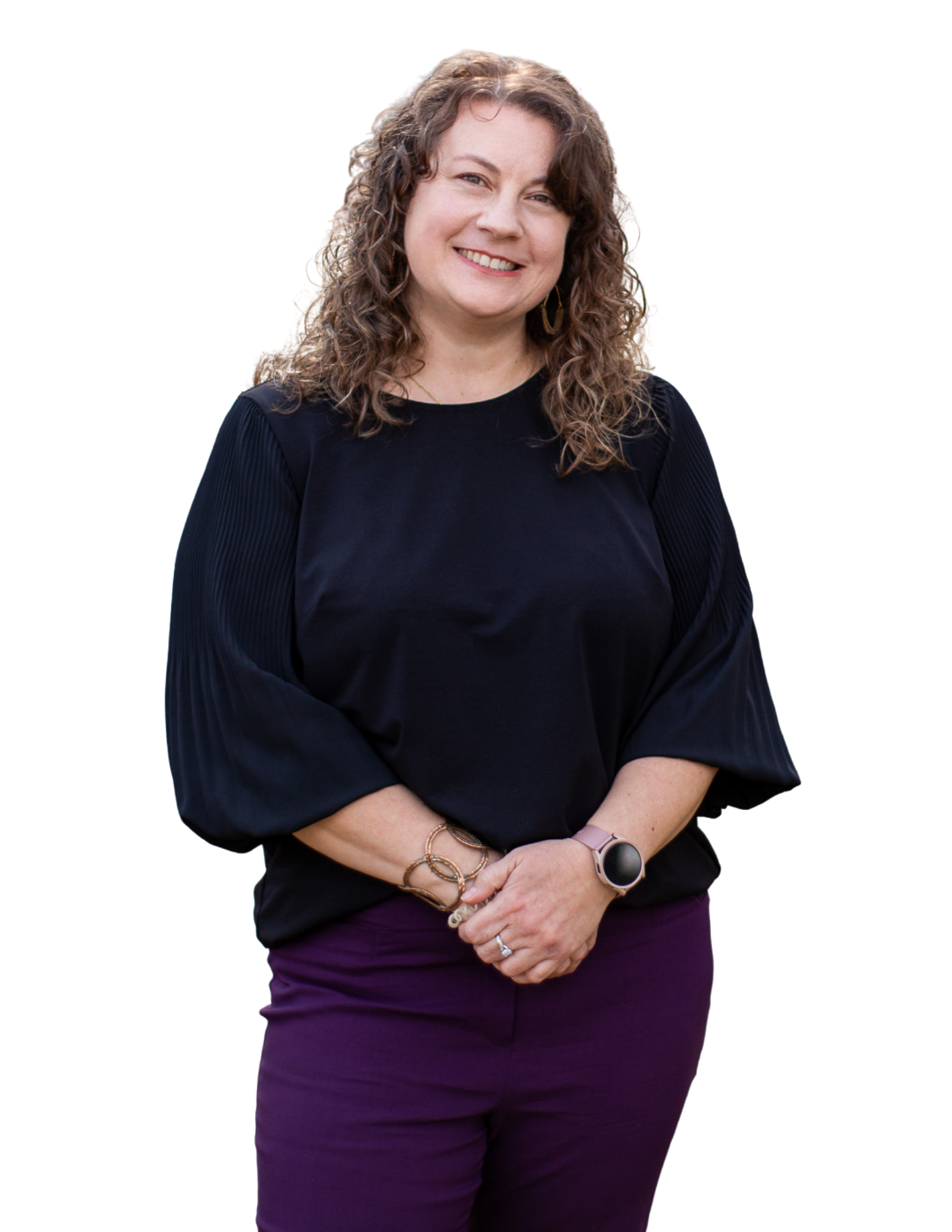 Smiling woman with curly brown hair, wearing a black blouse and purple pants, standing with her hands clasped in front of her.