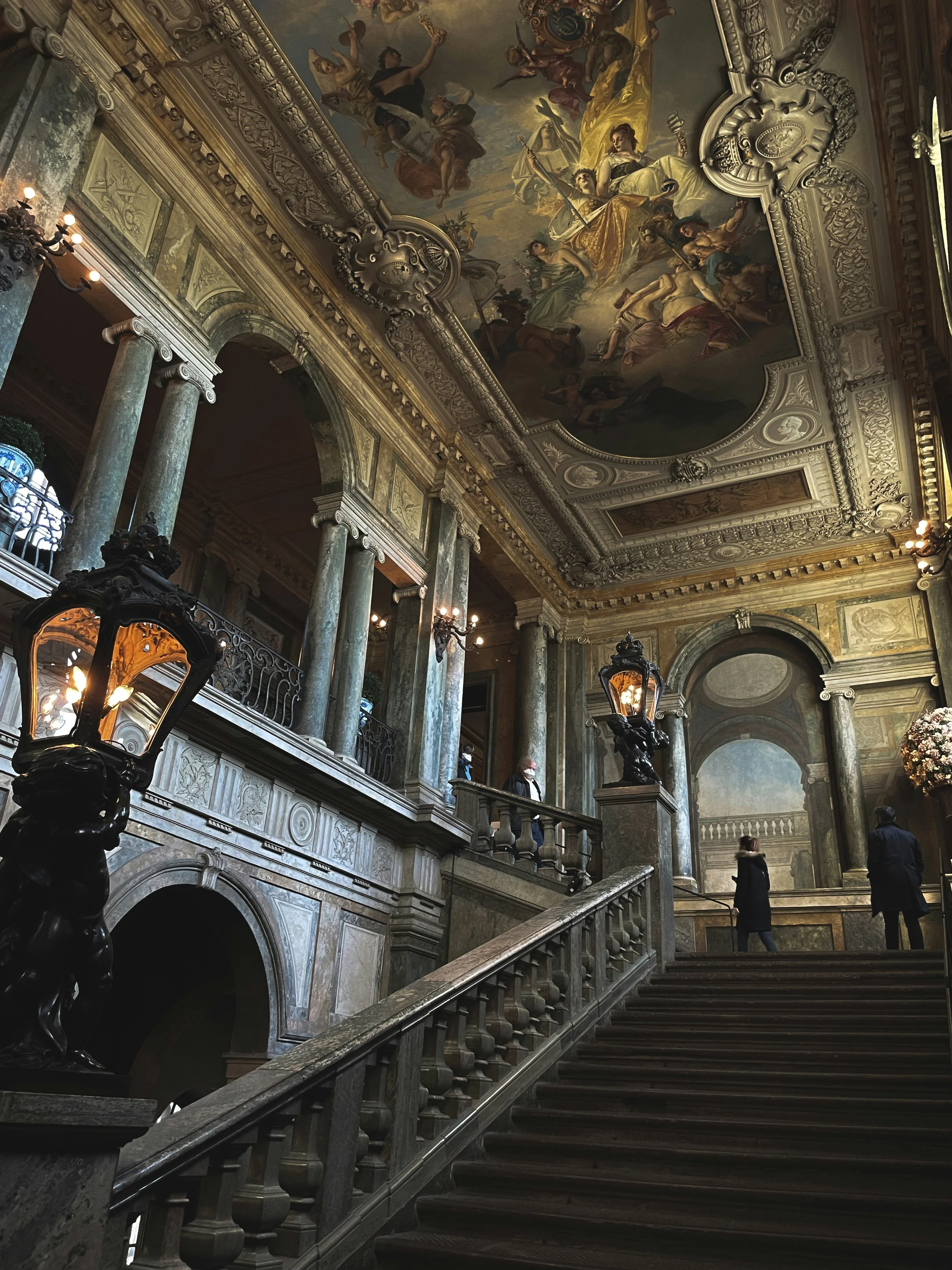 Interior of an ornate historic building with a grand staircase, marble columns, elaborate ceiling paintings, and vintage-style lamps.
