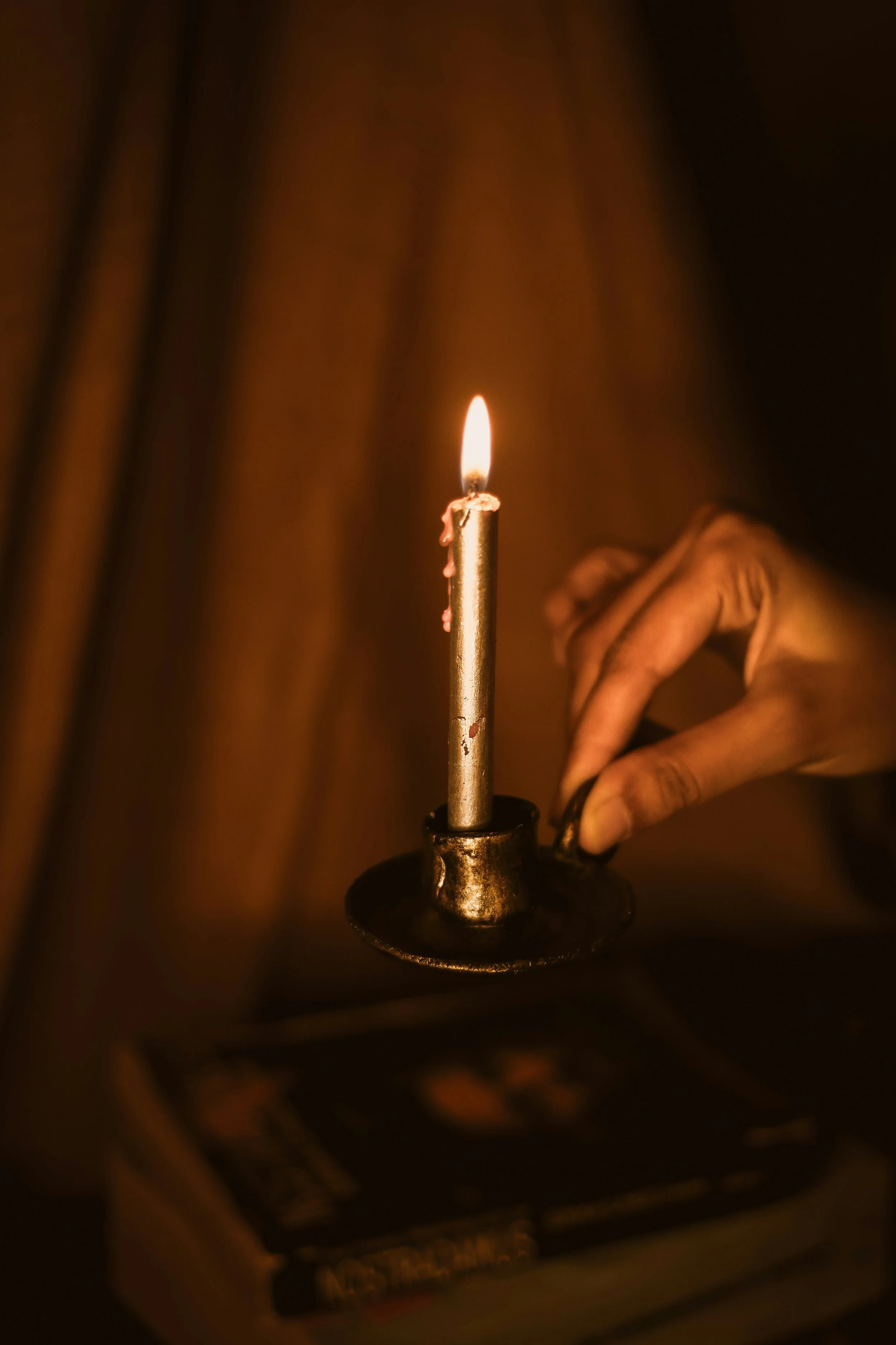 A hand holding a lit candle in a dark room, with a blurred book or DVD case on a table below.