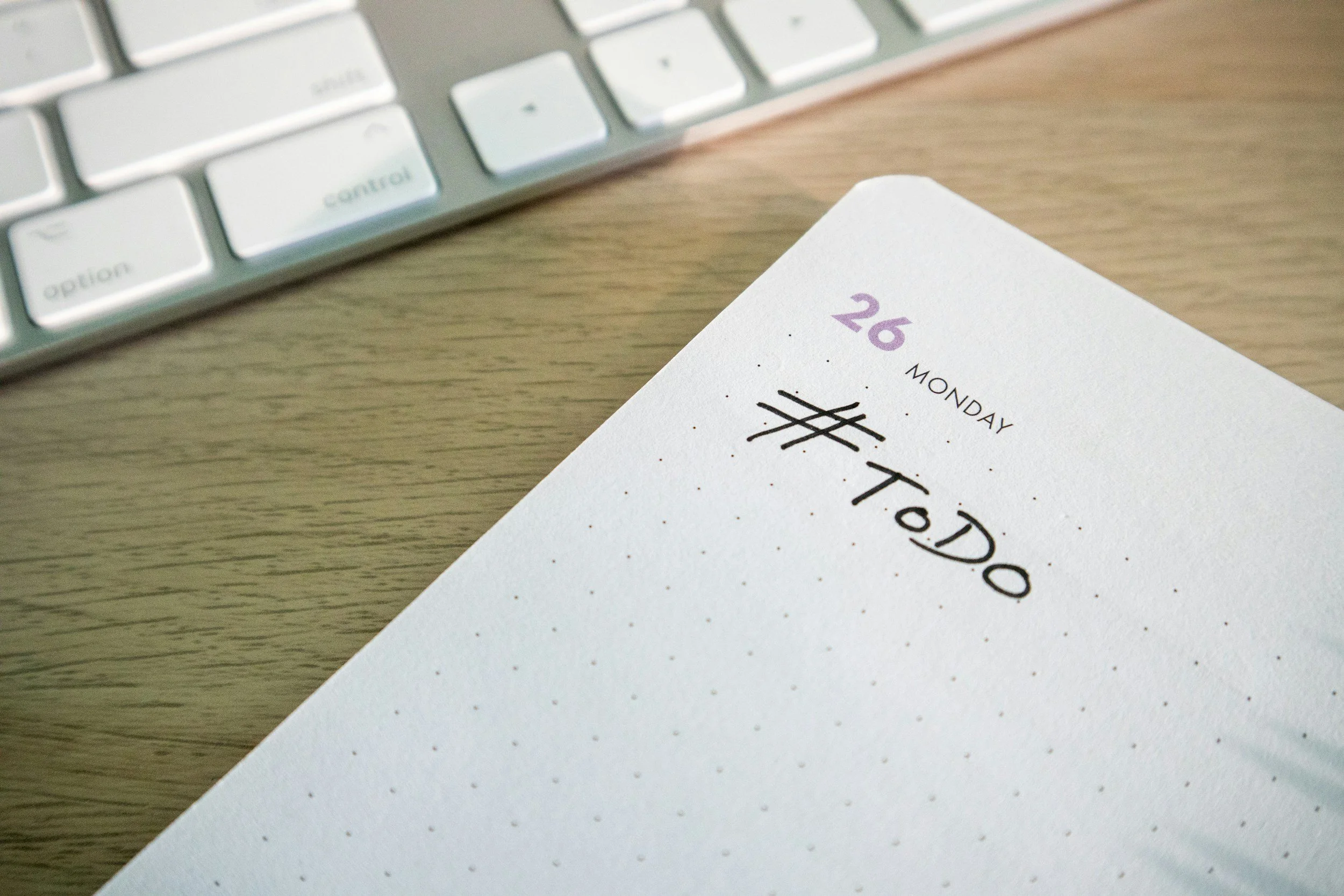 A printed planner page for Monday, the 26th, with the words 'To Do' handwritten and crossed out, on a wooden surface next to a white computer keyboard.
