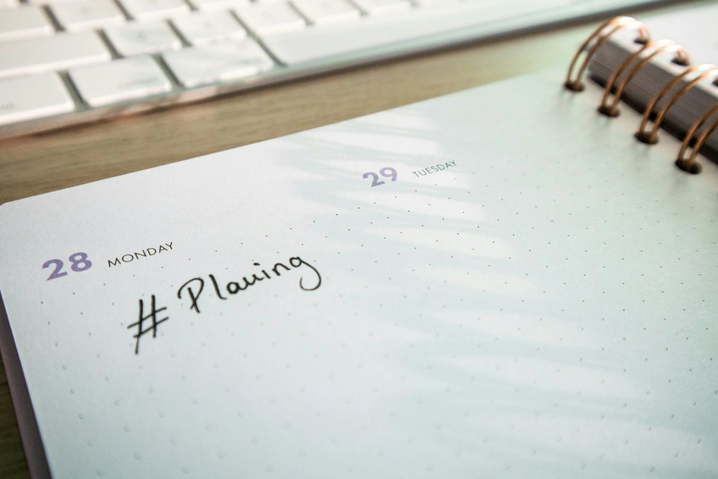 Open planner on a desk with a handwritten note that reads '# Planning', showing the dates Monday 28 and Tuesday 29. A keyboard and a spiral binding of the planner are also visible.