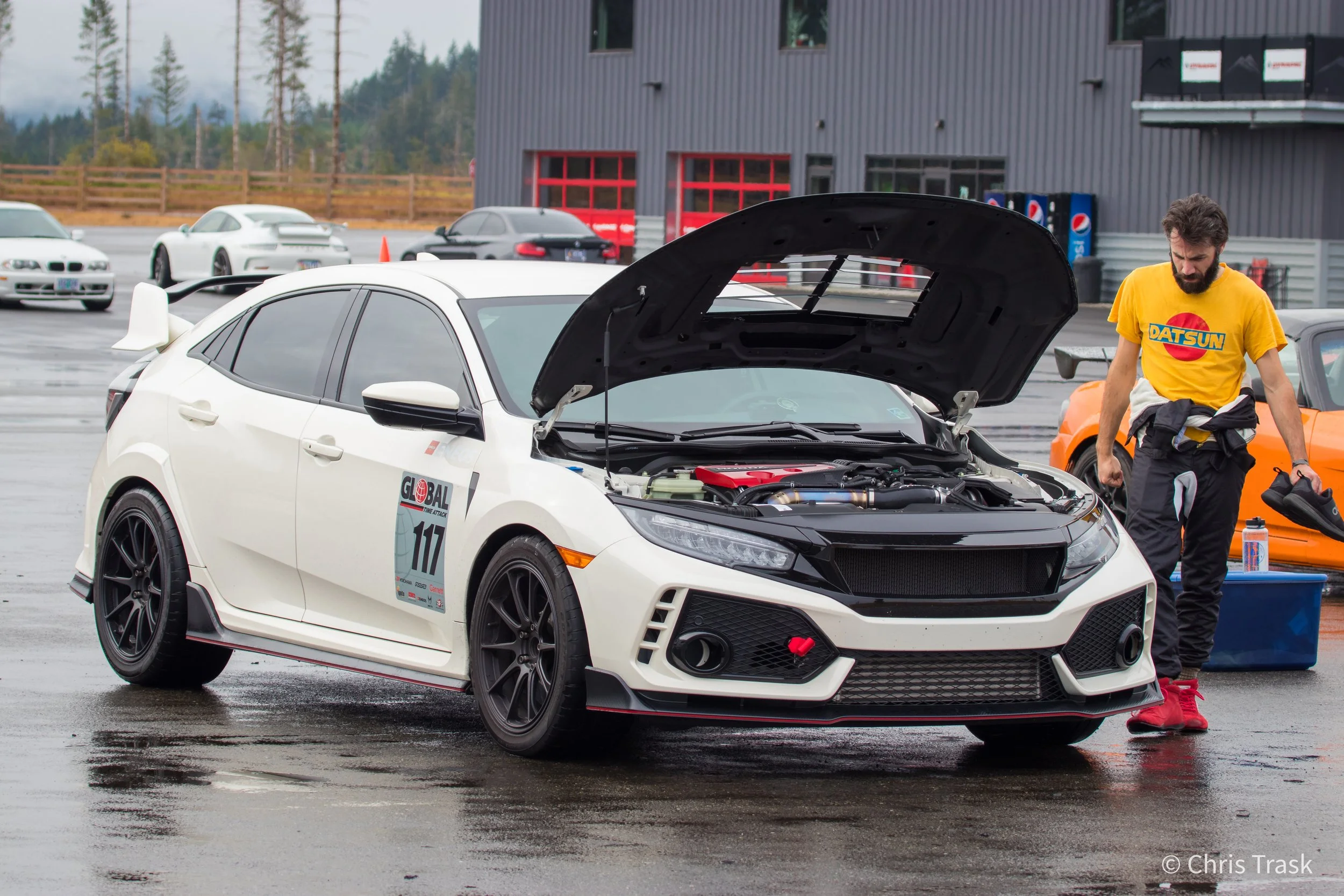A white sports car with its hood open at a racing event, with a man in a yellow Datsun t-shirt standing nearby, surrounded by other cars and racing building.