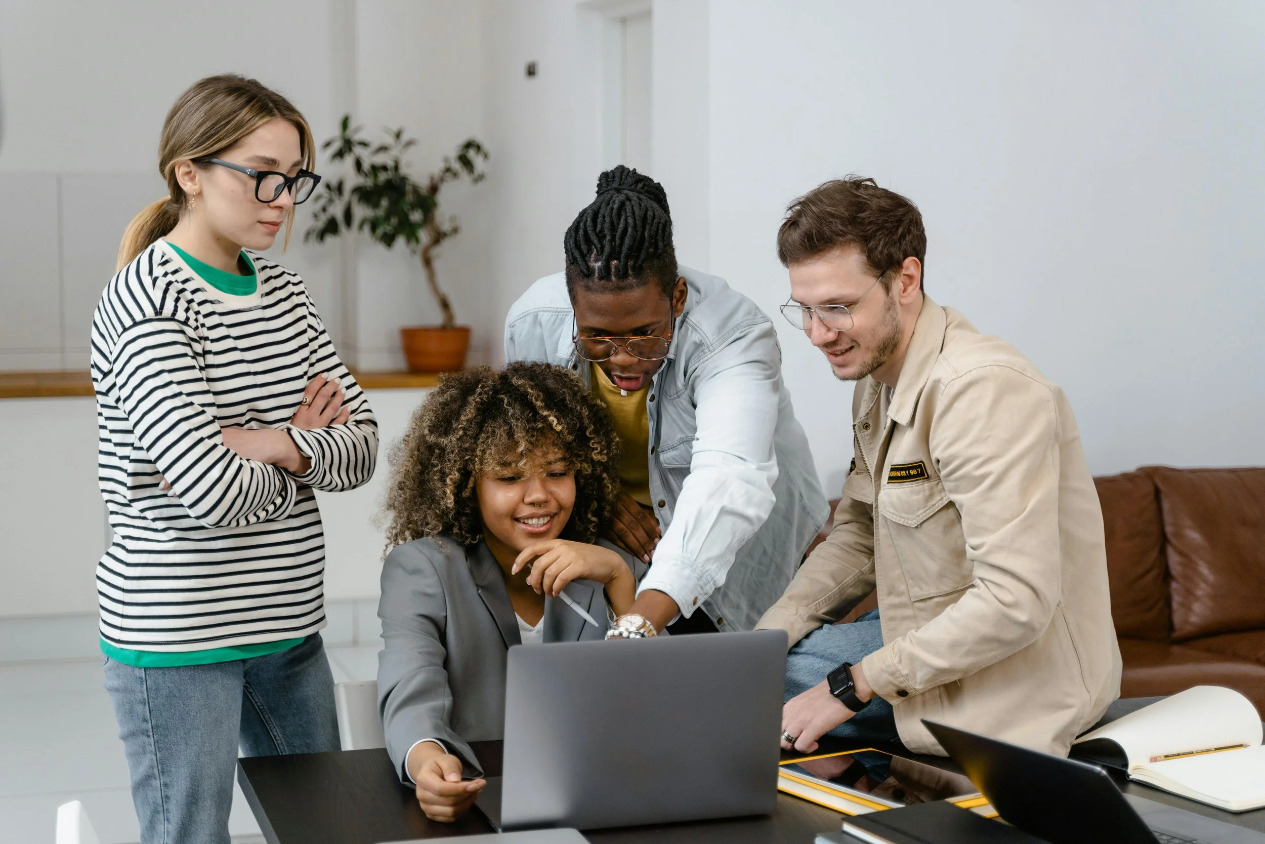 Group of four young adults gathered around a laptop, looking at the screen and smiling, in a modern office space.