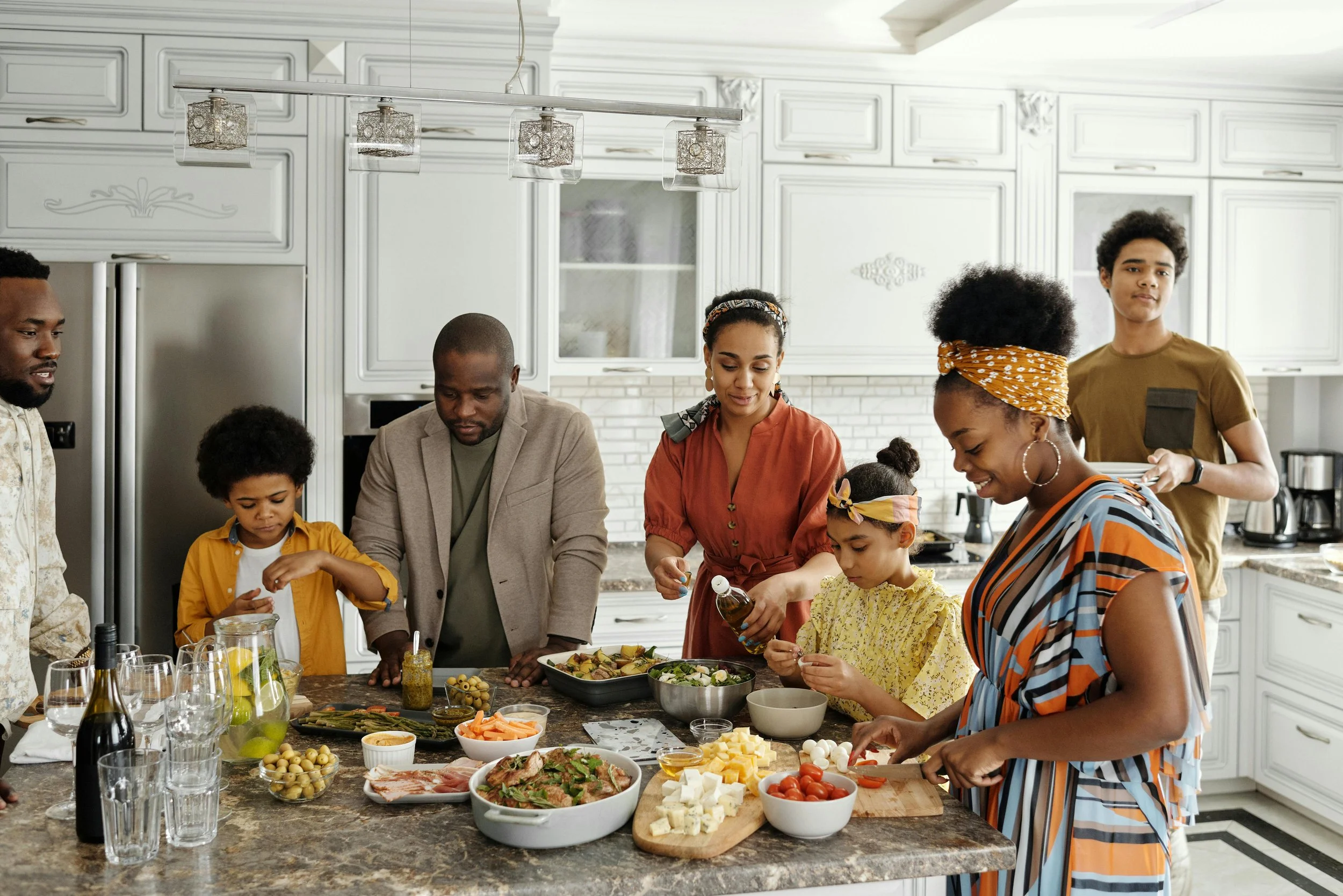 Family gathered around kitchen island preparing a meal with various ingredients like vegetables, cheese, and bread.