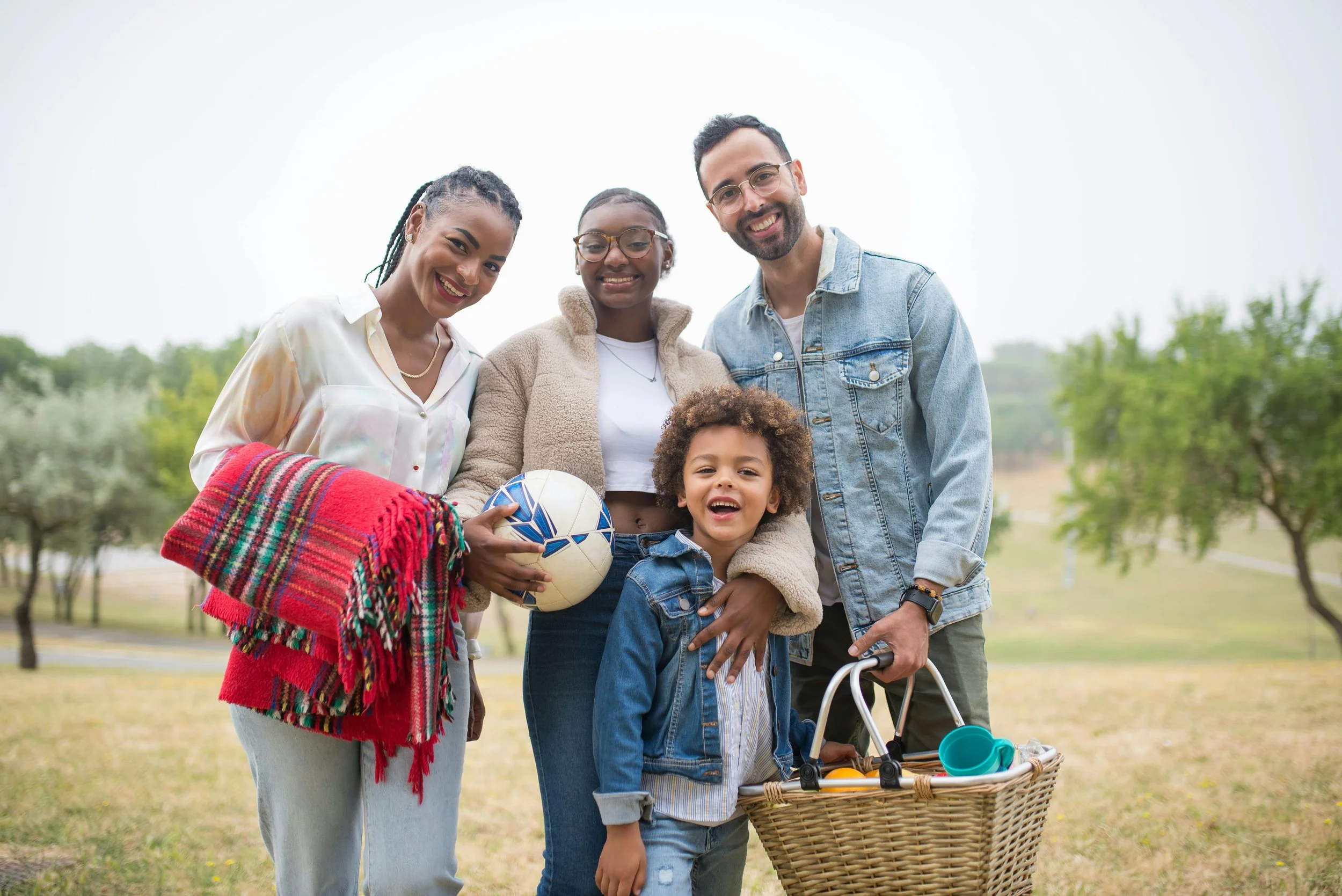 A diverse group of five people, including two women, one man, a young girl, and a young boy, smiling and standing outdoors in a park with trees in the background. The girl is holding a soccer ball, and the boy is holding a bicycle basket with toys.