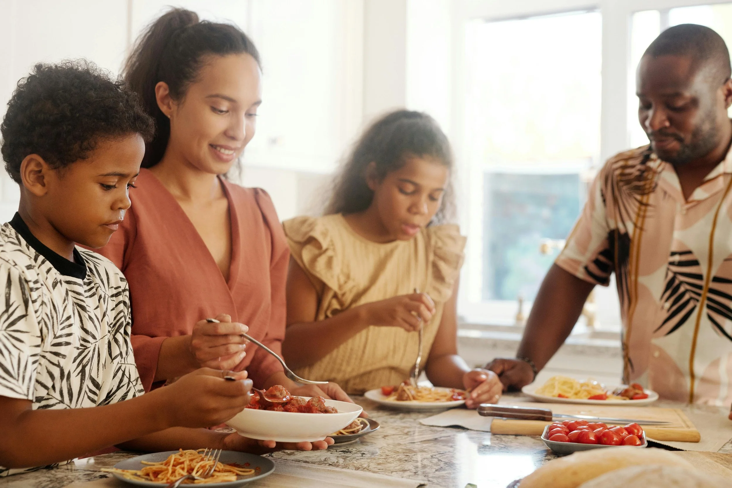 Family of four preparing and serving pasta and tomatoes in a bright kitchen.