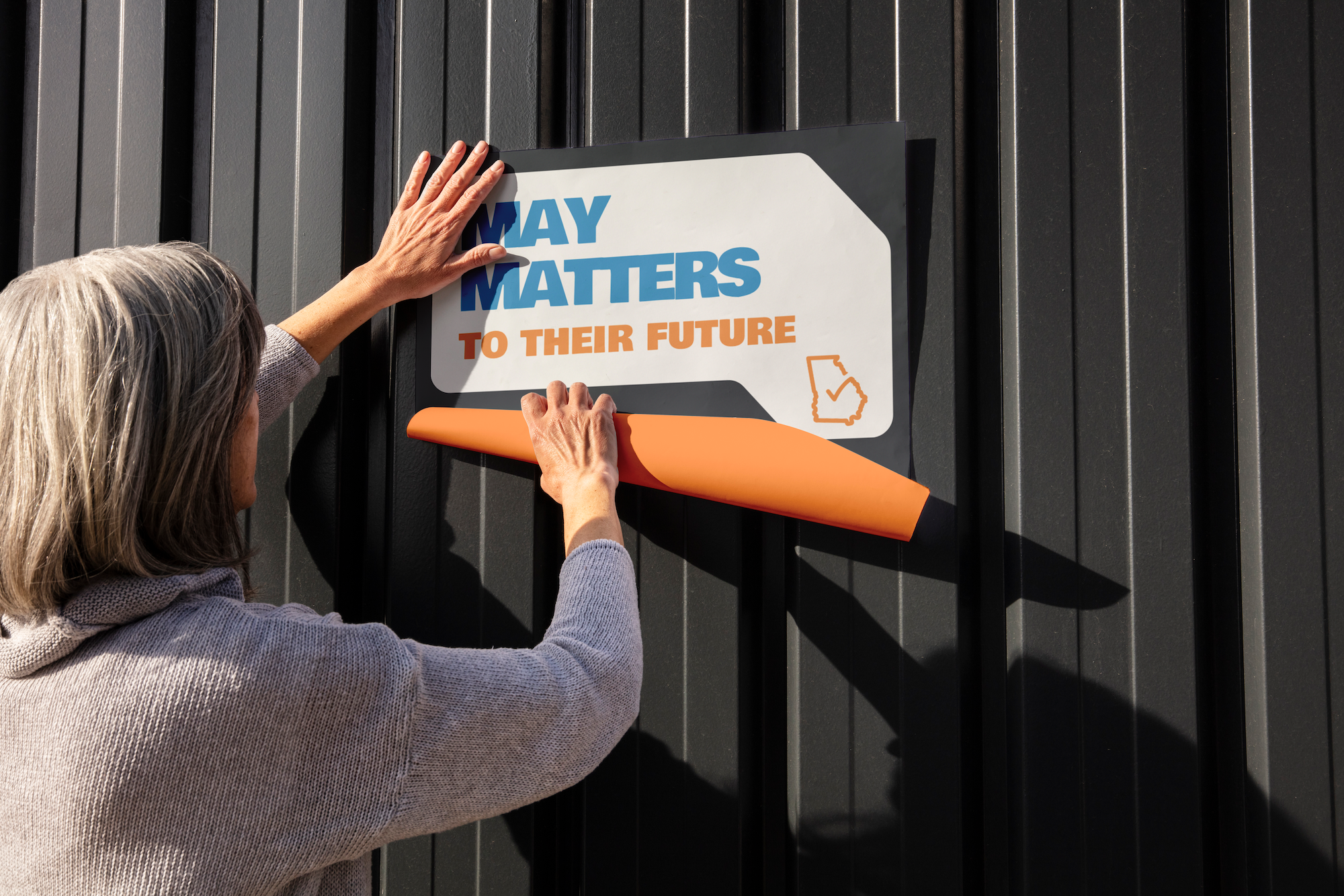 A woman peels off a poster that reads "MAY MATTERS TO THEIR FUTURE" on a black wall. This demonstrates a poster design for the May Matters nonpartisan community civic engagement initiative ahead of the May primary in Georgia.