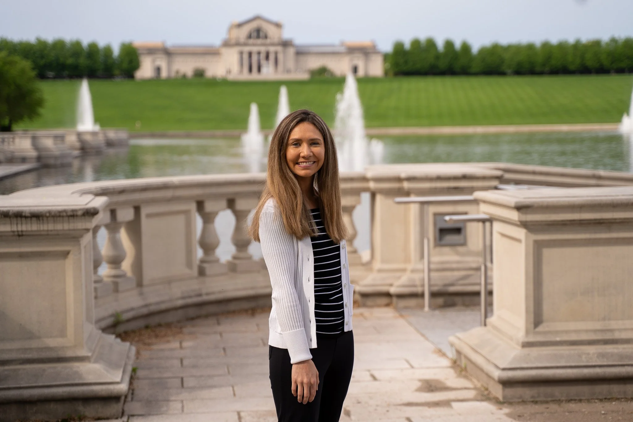 A woman with long brown hair smiling at the camera, standing on a stone balcony overlooking a pond with fountains and green grass, with a large historic building in the background.