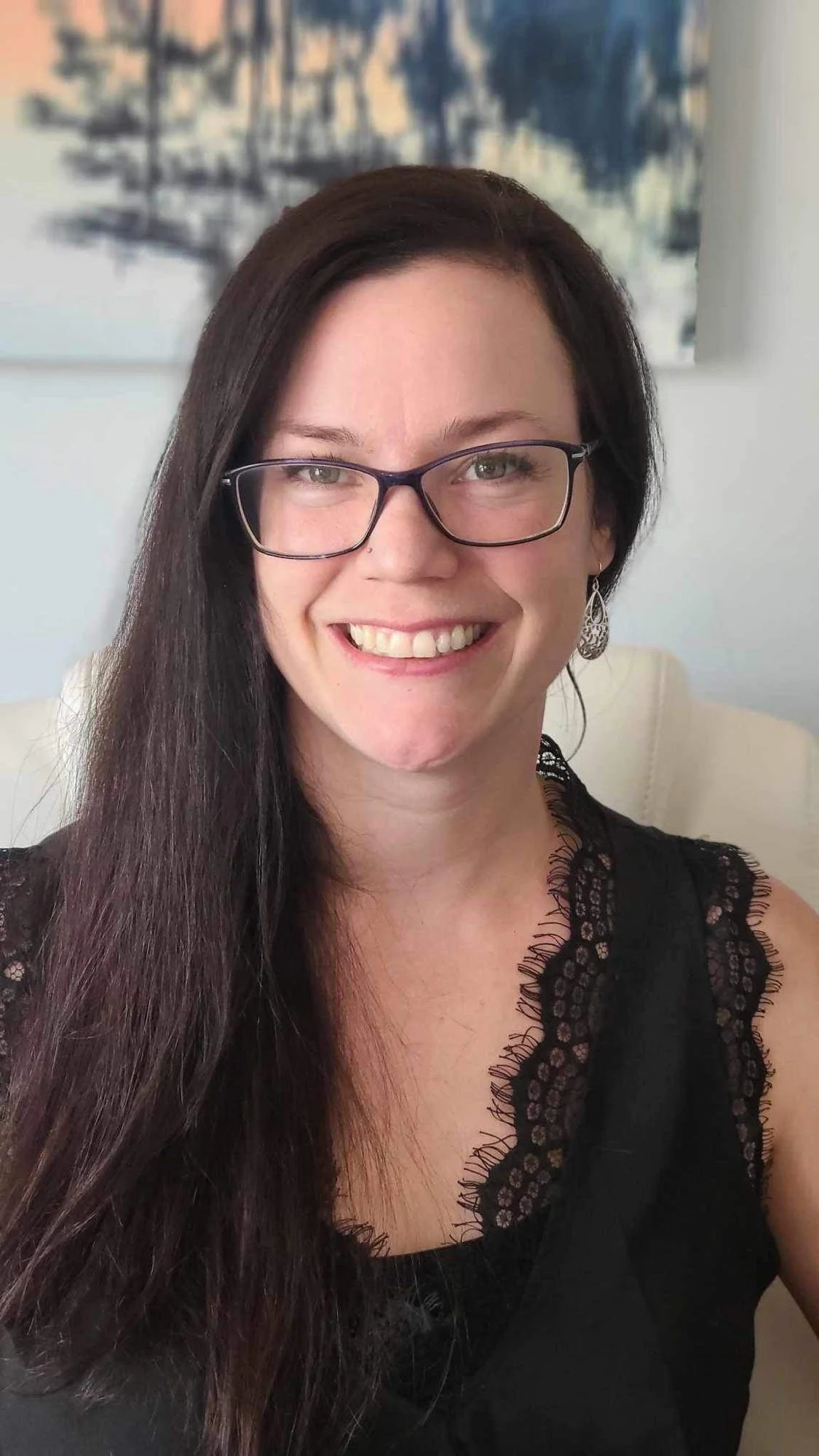 A woman with long dark hair, glasses, and earrings, smiling while sitting in a room with a modern painting in the background.