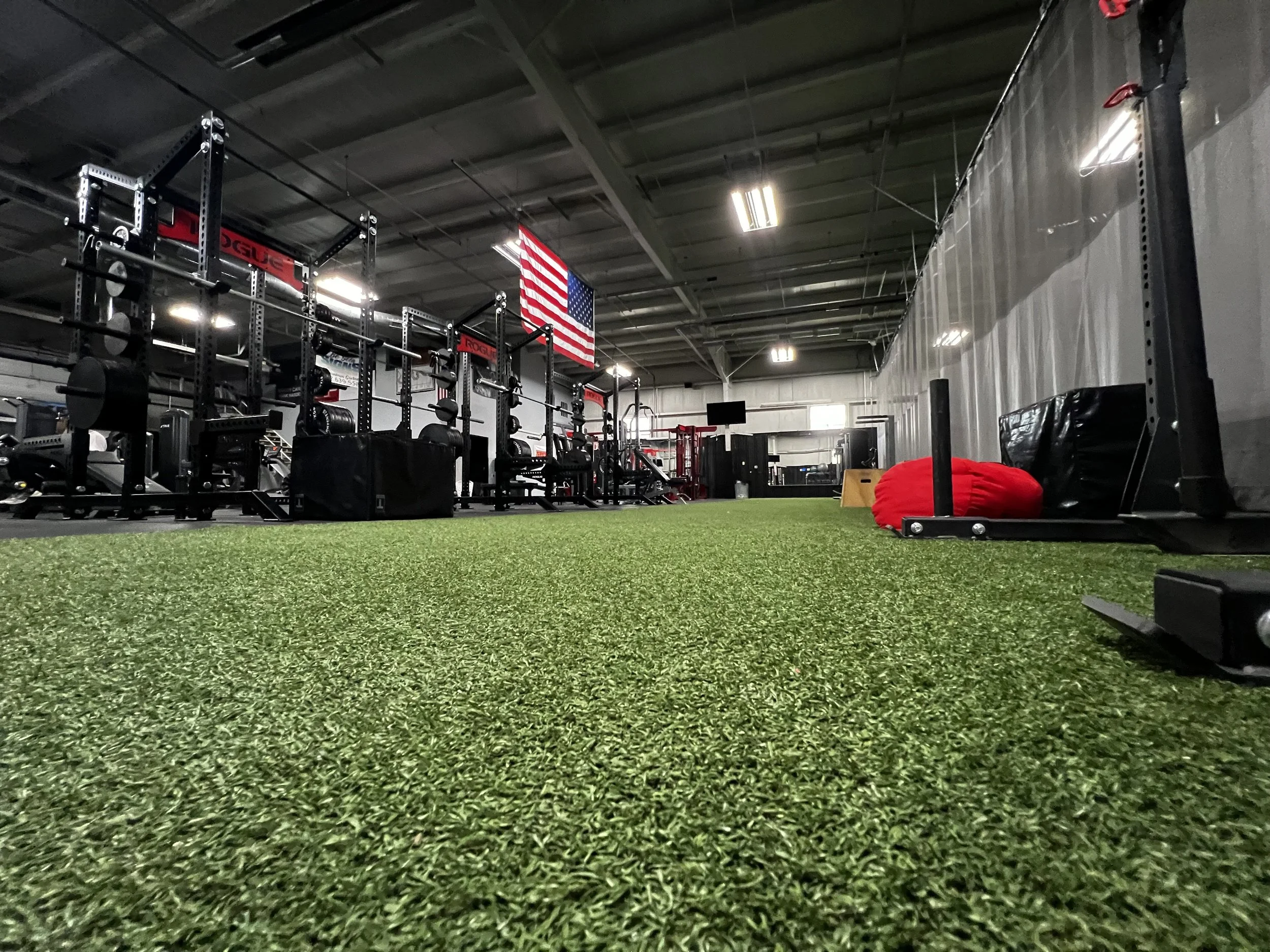 Interior view of a gym with artificial turf flooring, weightlifting equipment, an American flag, and red workout gear.
