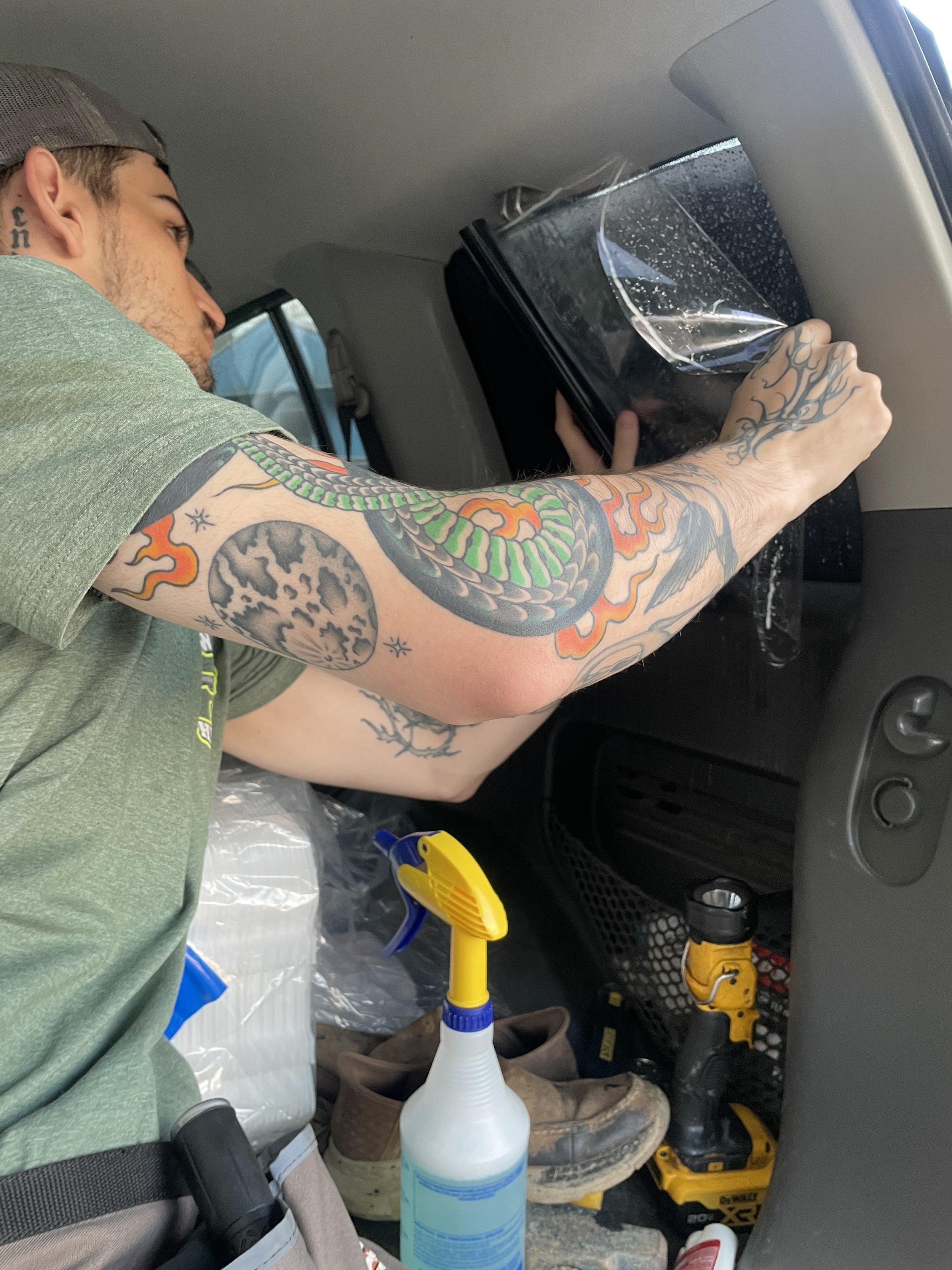 A man with tattoos working inside a vehicle, pulling the liner off of window tint. The interior contains various tools and supplies, including a spray bottle.