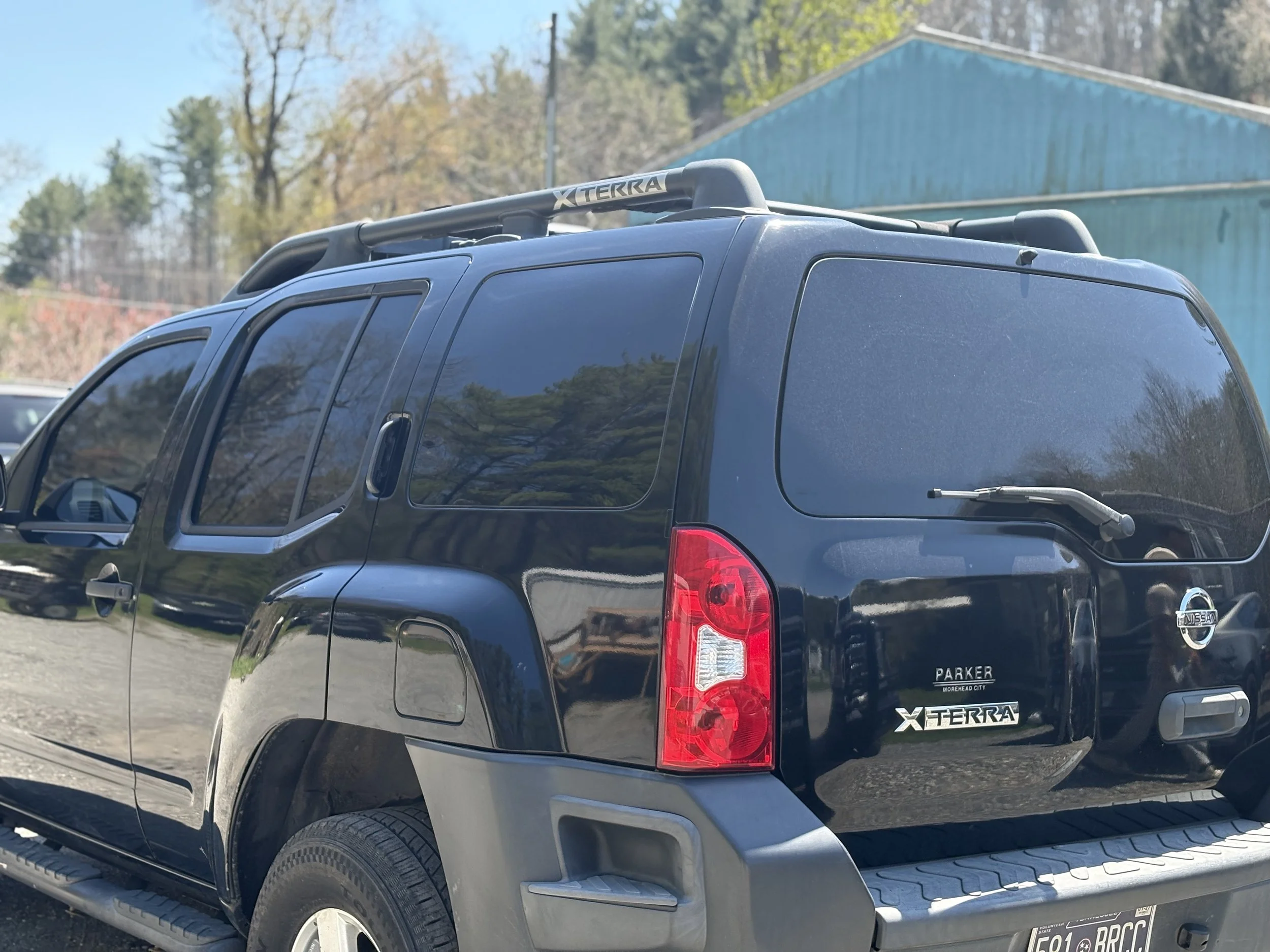 A freshly tinted, black Nissan Xterra SUV parked outdoors with trees and a teal building in the background.
