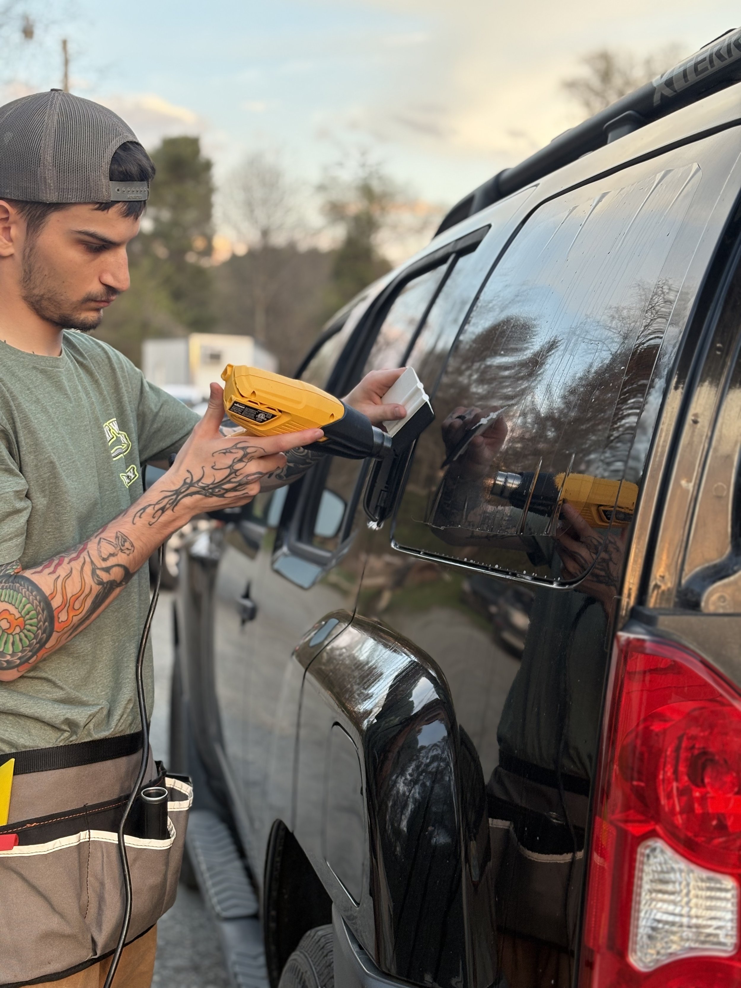 A man wearing a gray cap and a green T-shirt with tattoos on his arms holds a heat gun against the side window of a black SUV. The scene is outdoors with trees and a partly cloudy sky in the background.