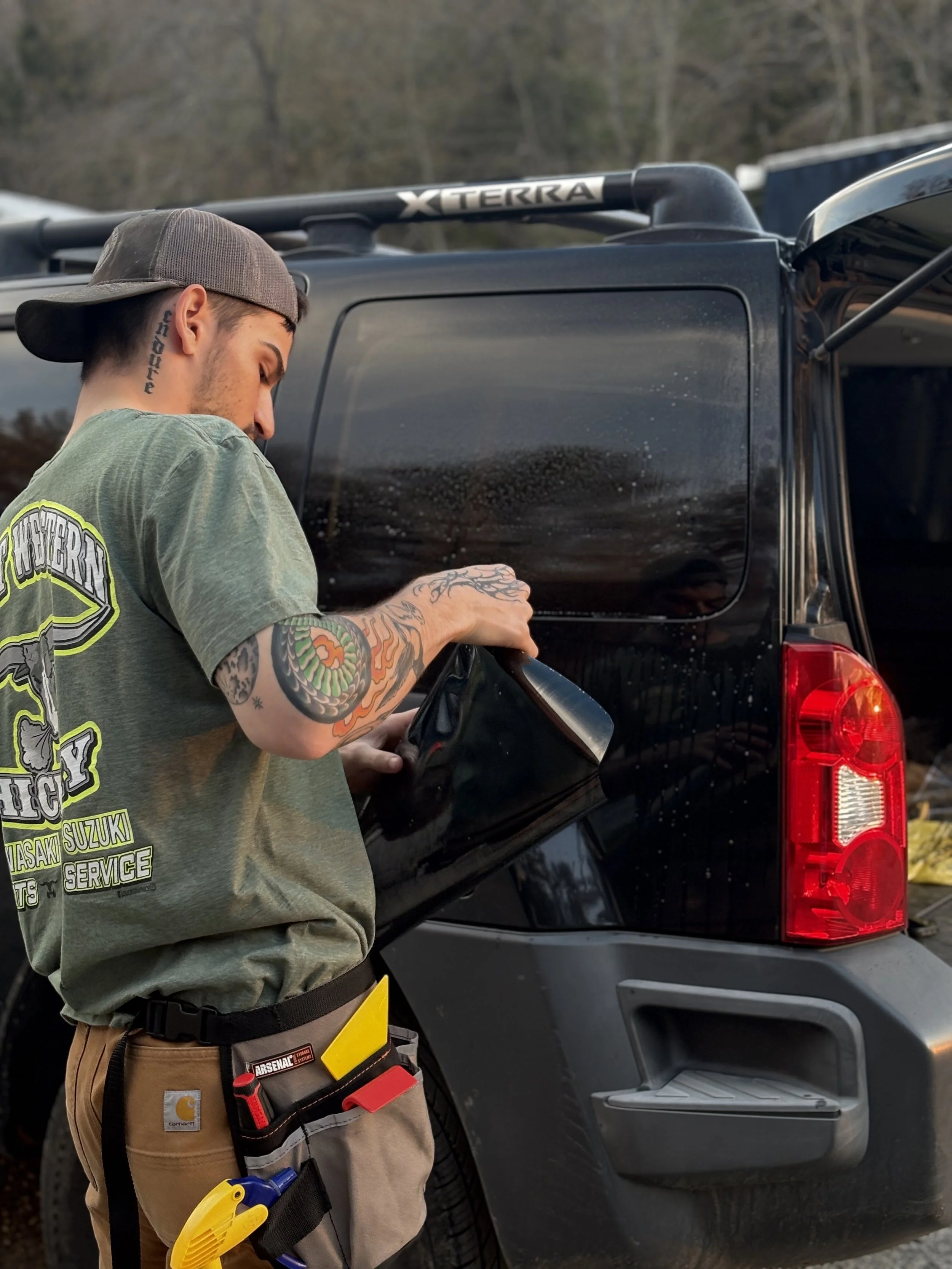 A man wearing a backwards baseball cap is working on the side of a black SUV. He has a tool belt with various tools around his waist.