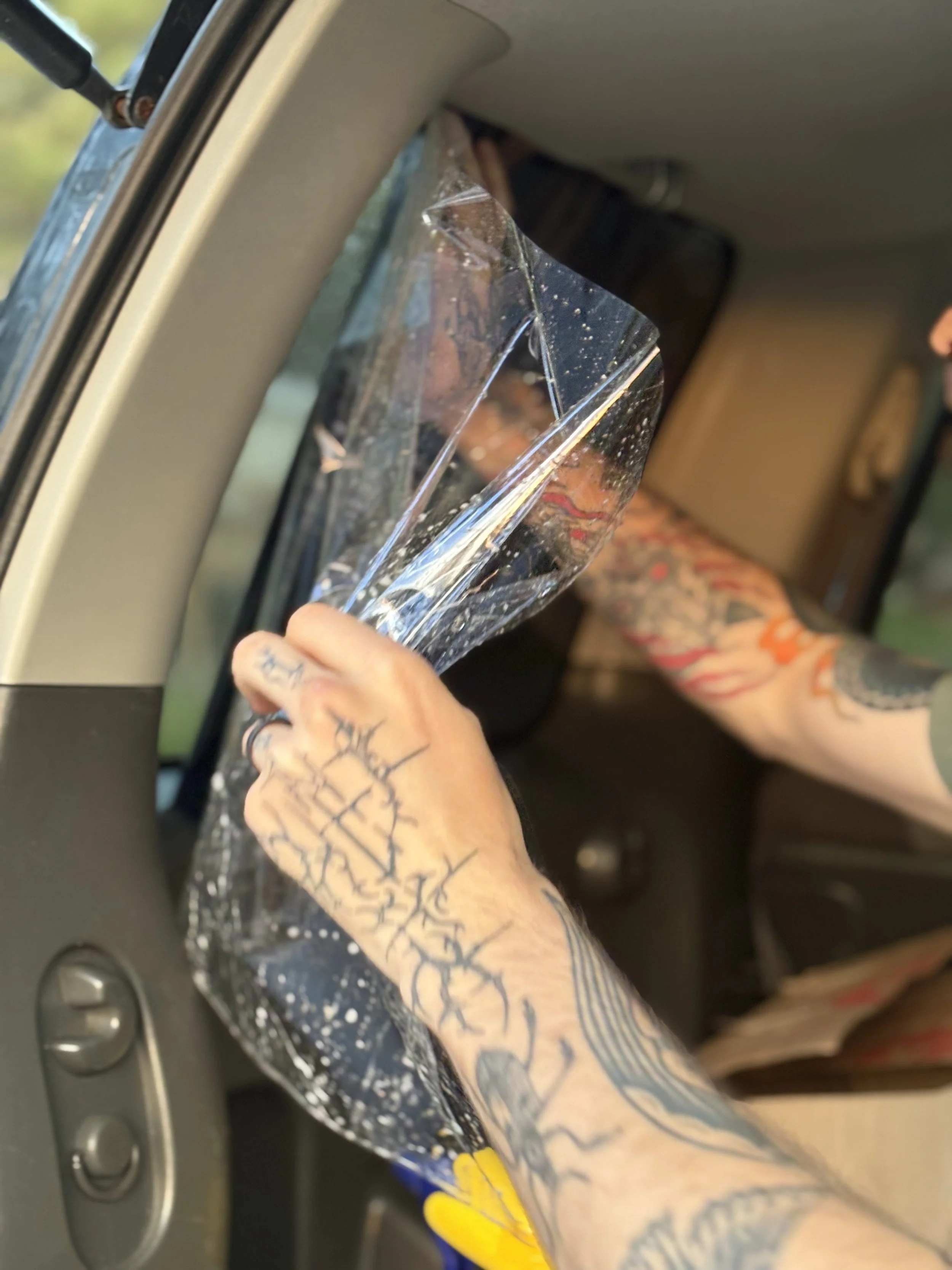 Close-up of a person's tattooed arm installing a piece of tint on the inside of a vehicle. The vehicle's dashboard and window are visible.