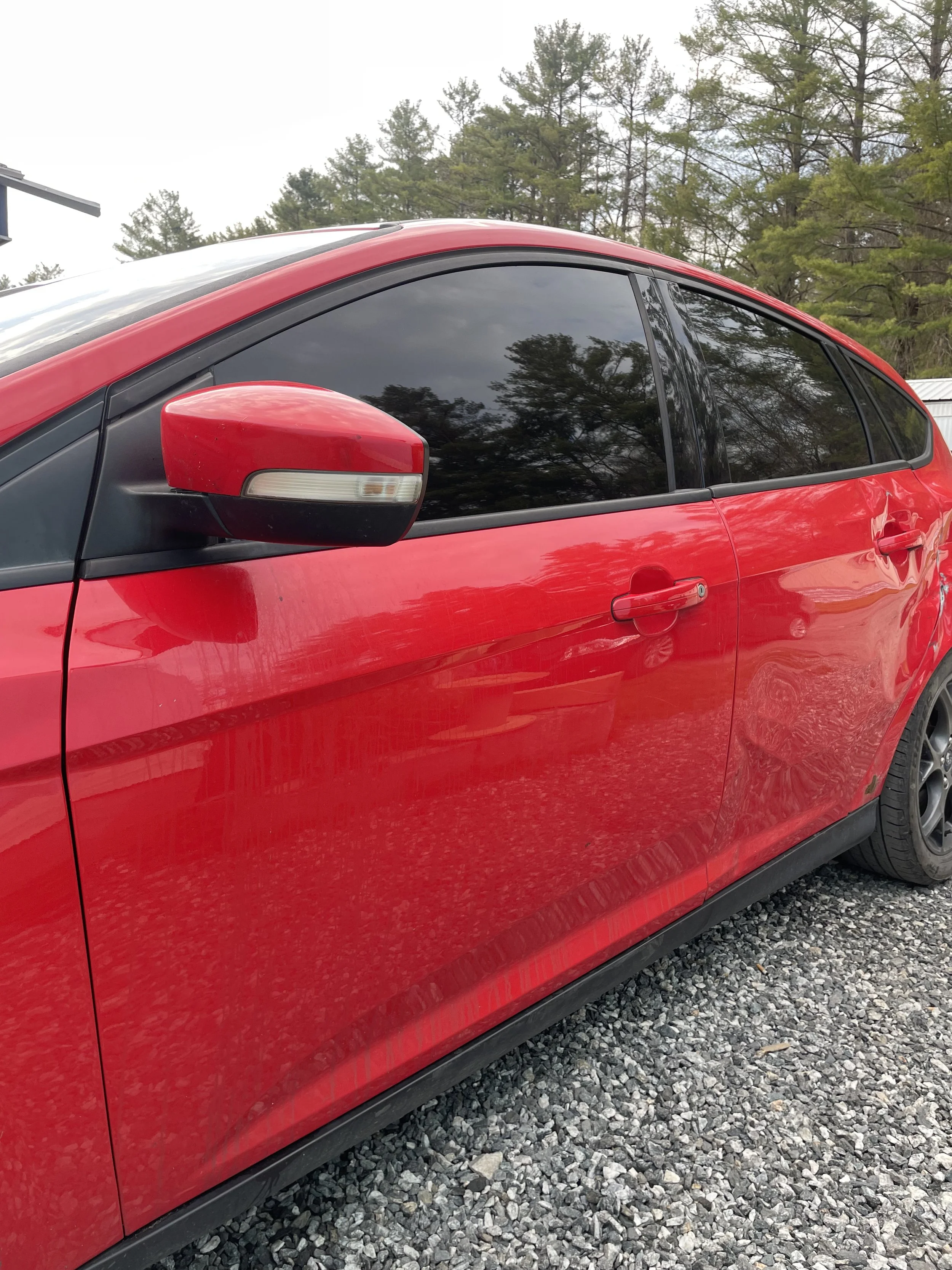 A tinted, red Ford Focus parked on a gravel lot with trees and overcast sky in the background.