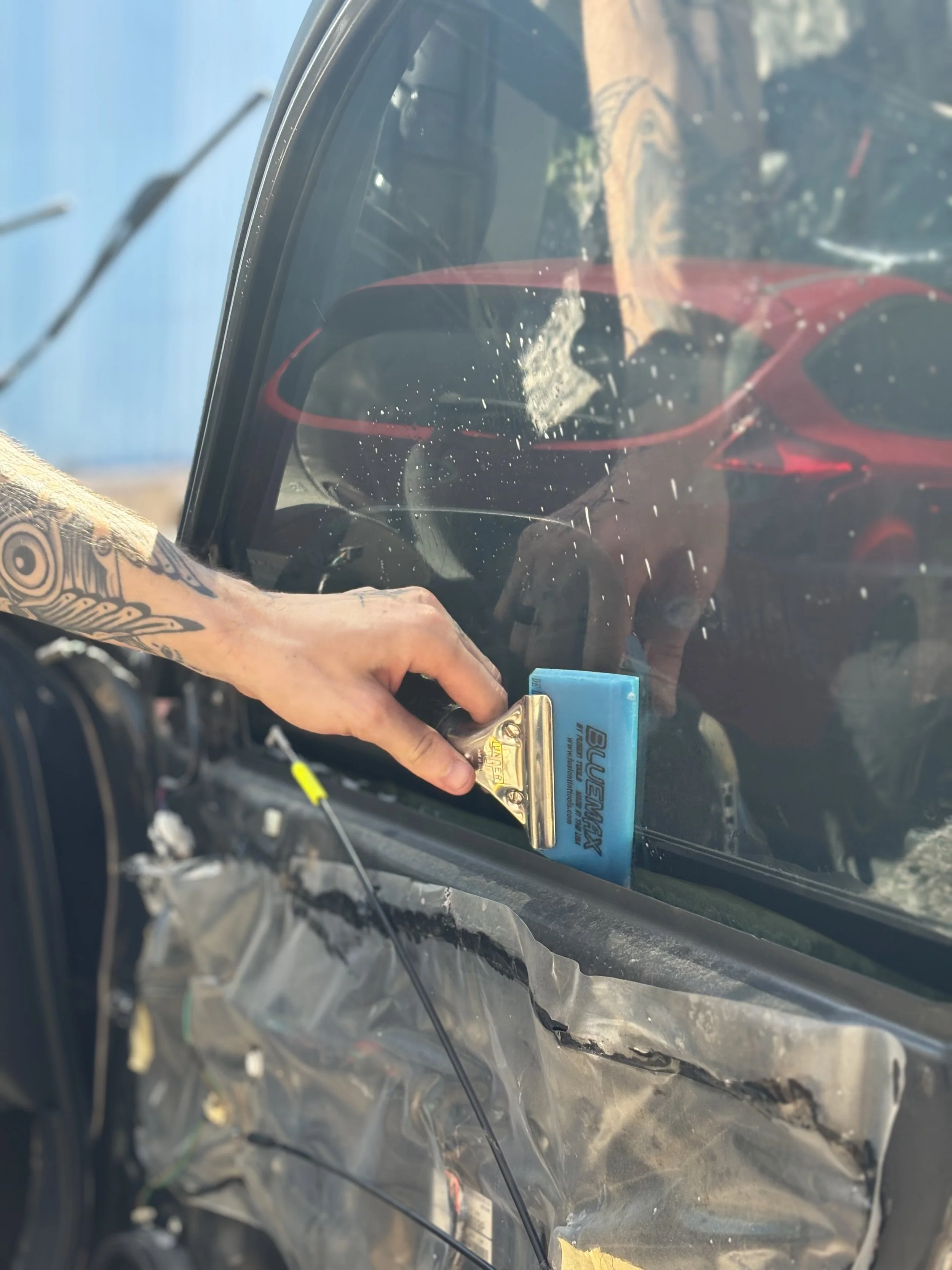 A person with a tattoo on their arm is holding a metal tool with a yellow label and blue plastic part, working inside a car door, with the window slightly open. The car's interior and some wires are visible, with a blurry background outside the car.