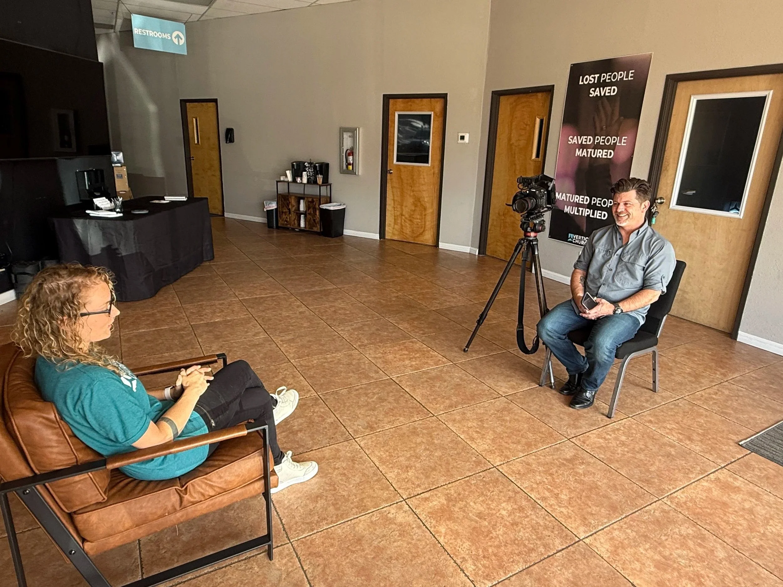 A woman with curly hair wearing glasses and a teal shirt sitting on a brown leather chair, looking at her phone. In the background, a man is sitting on a chair, smiling, with a video camera on a tripod pointed at him. The room has brown tiled flooring, with four wooden doors on the back wall and a large poster with motivational phrases about saved and matured people. There is a small table with coffee supplies near the doors, and an overhead blue sign indicating the restrooms.