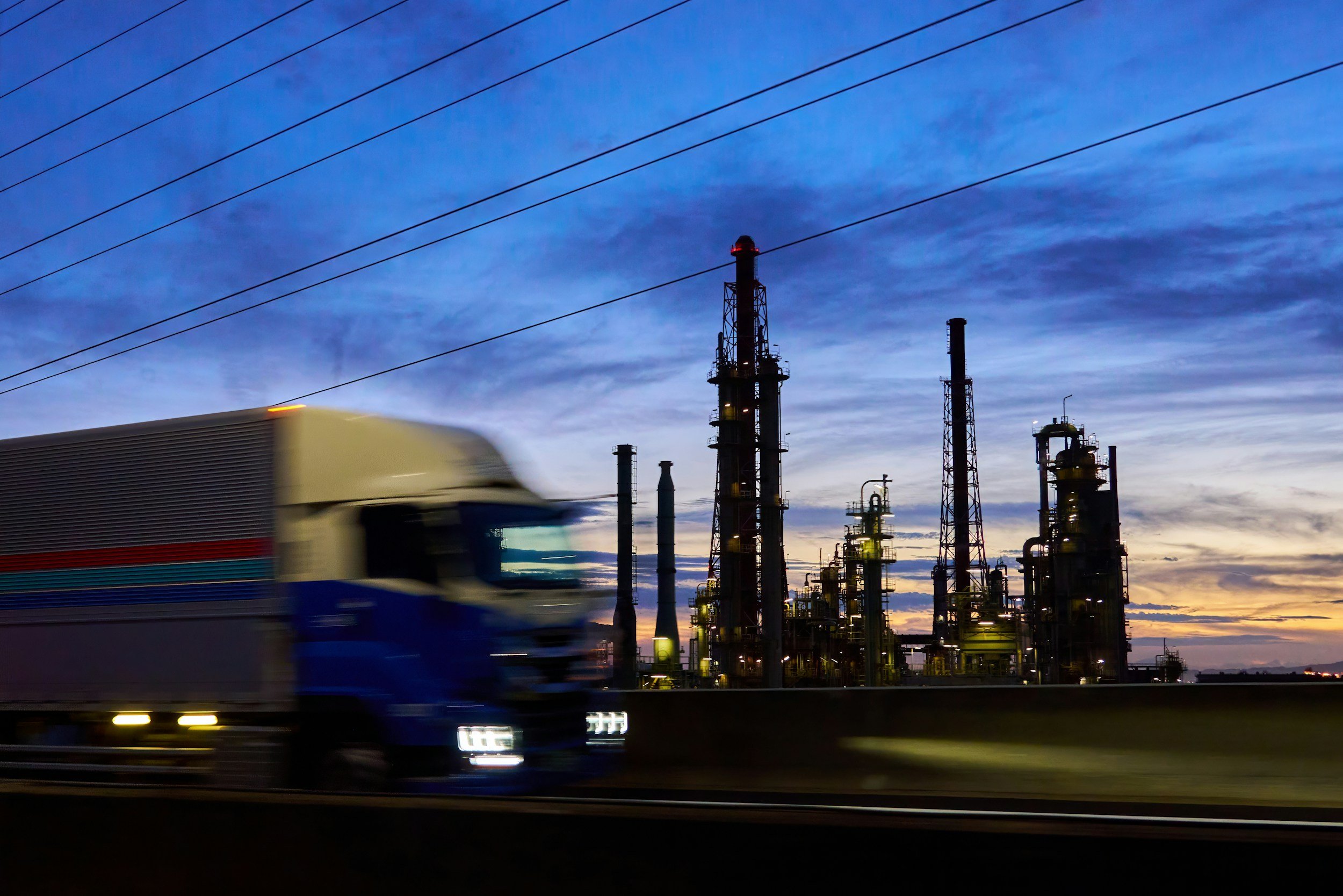 A blurred blue and white truck moving past an industrial refinery with tall structures, pipes, and towers against a blue sky at dusk.