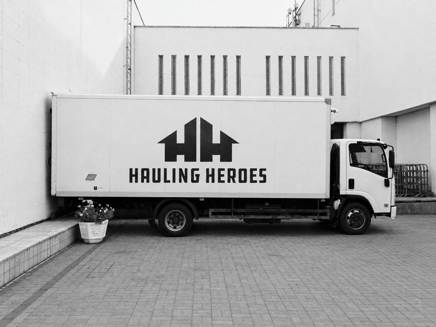 A white moving truck with a logo featuring two black house silhouettes and the text 'Hauling Heroes' parked on a paved area near a white building with a small flowerpot in front.