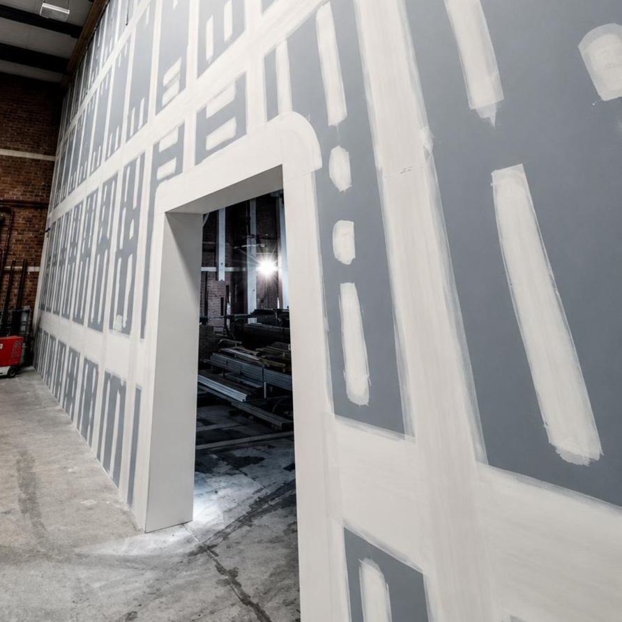 Indoor construction site with a partially painted gray wall featuring large white outlined lettering and geometric shapes, with an open doorway revealing construction materials inside.