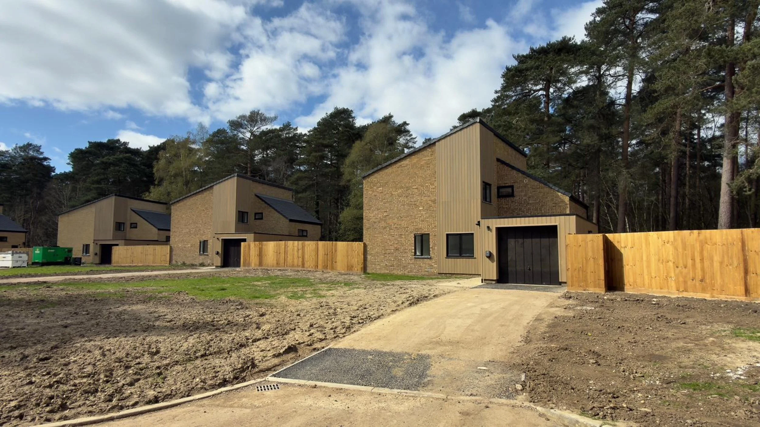 Three modern houses under construction with brown brick and siding, surrounded by a dirt and grass landscape, and dense trees in the background under a partly cloudy sky.