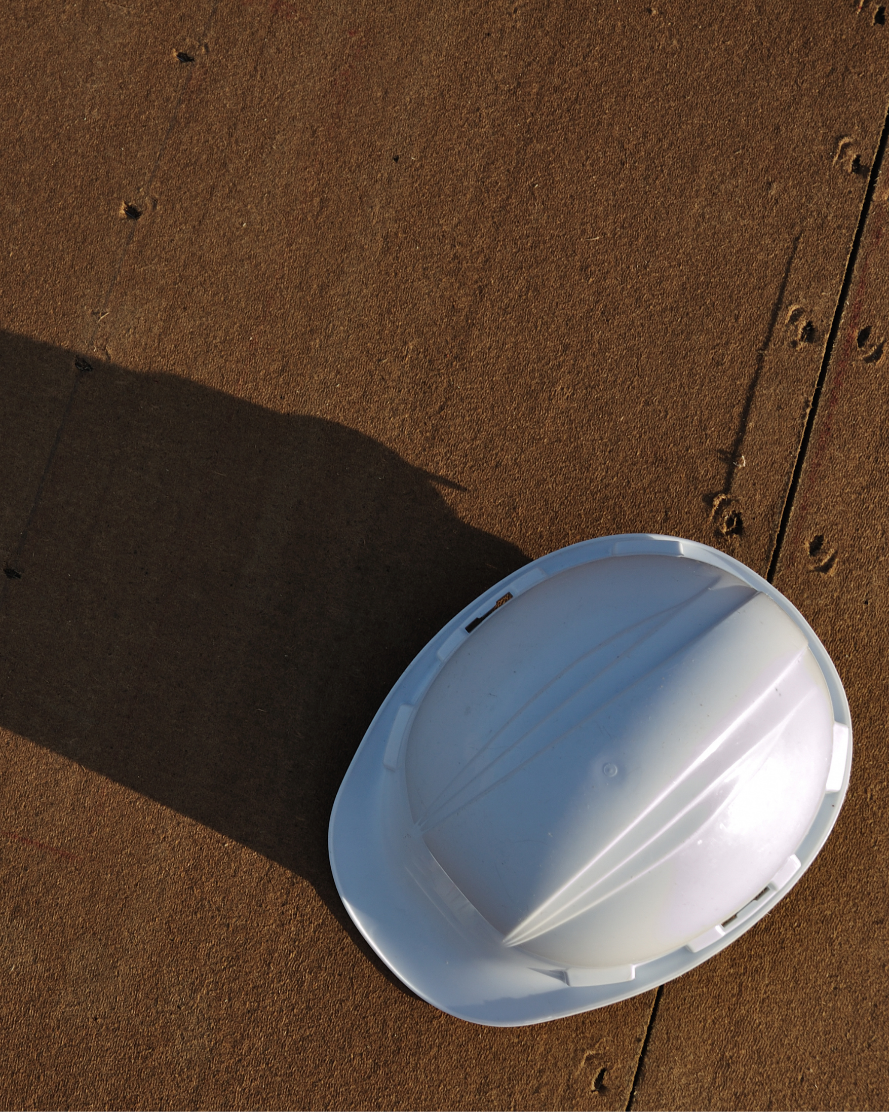A white hard hat casting a long shadow on a reddish-brown surface, possibly a wooden or metal floor.