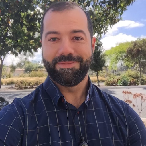 A man with a beard and short hair smiling outdoors, wearing a dark plaid shirt, with trees and a parking lot in the background.