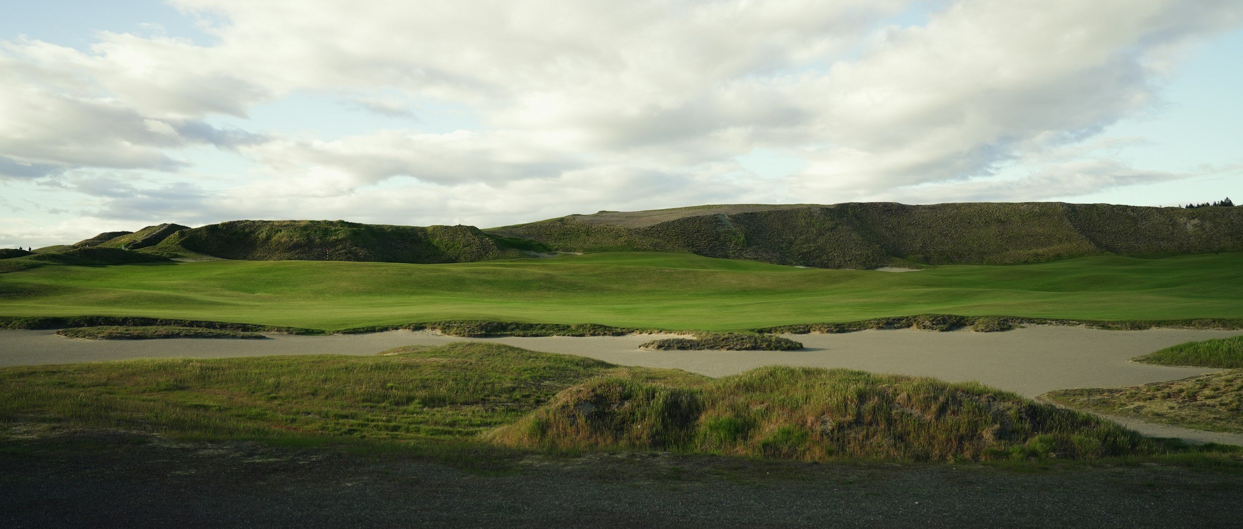 A scenic view of a golf course with lush green grass, sand bunkers, and rolling hills under a partly cloudy sky.