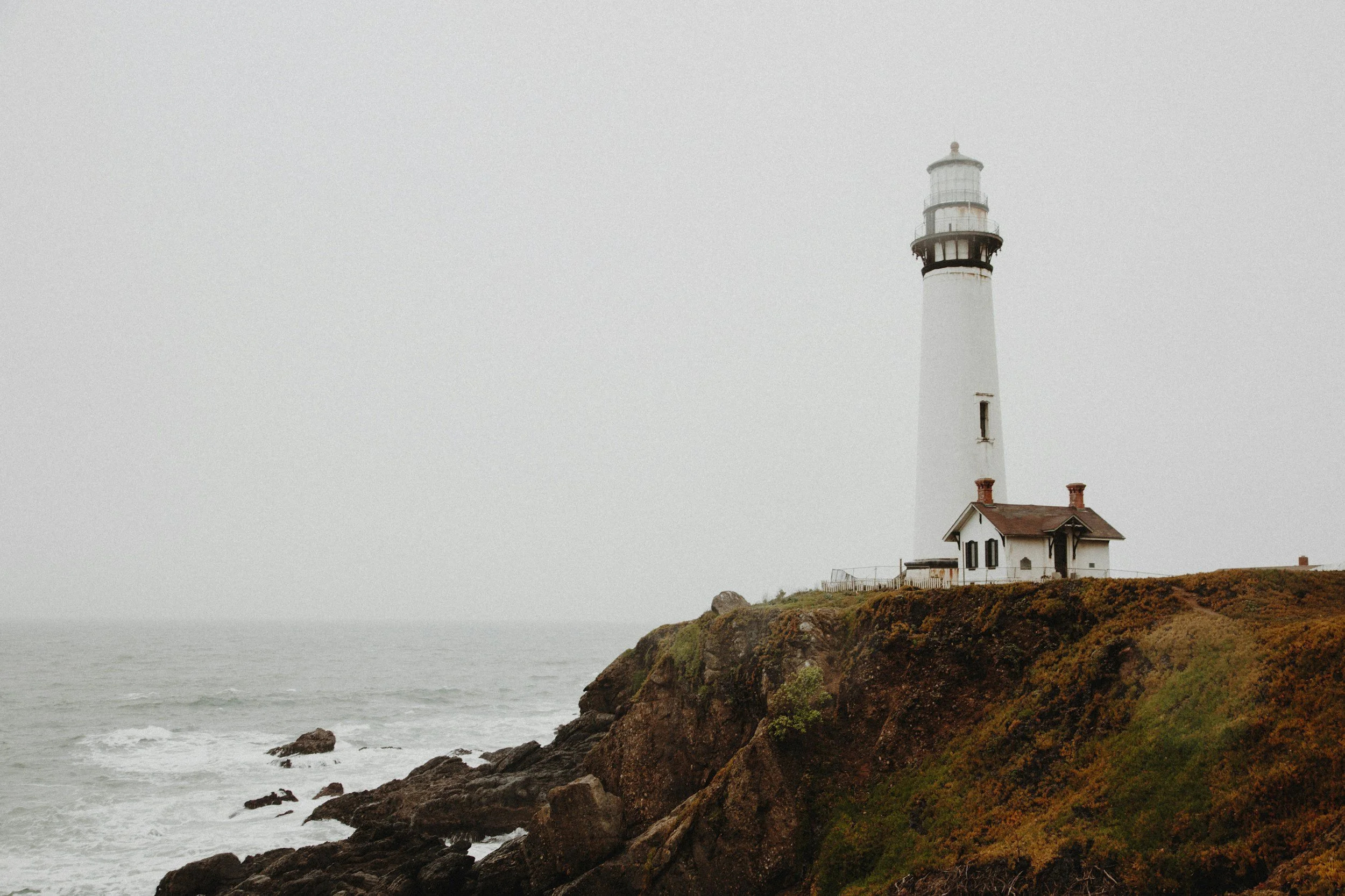 A lighthouse on a rocky cliff overlooking the ocean on a foggy day.