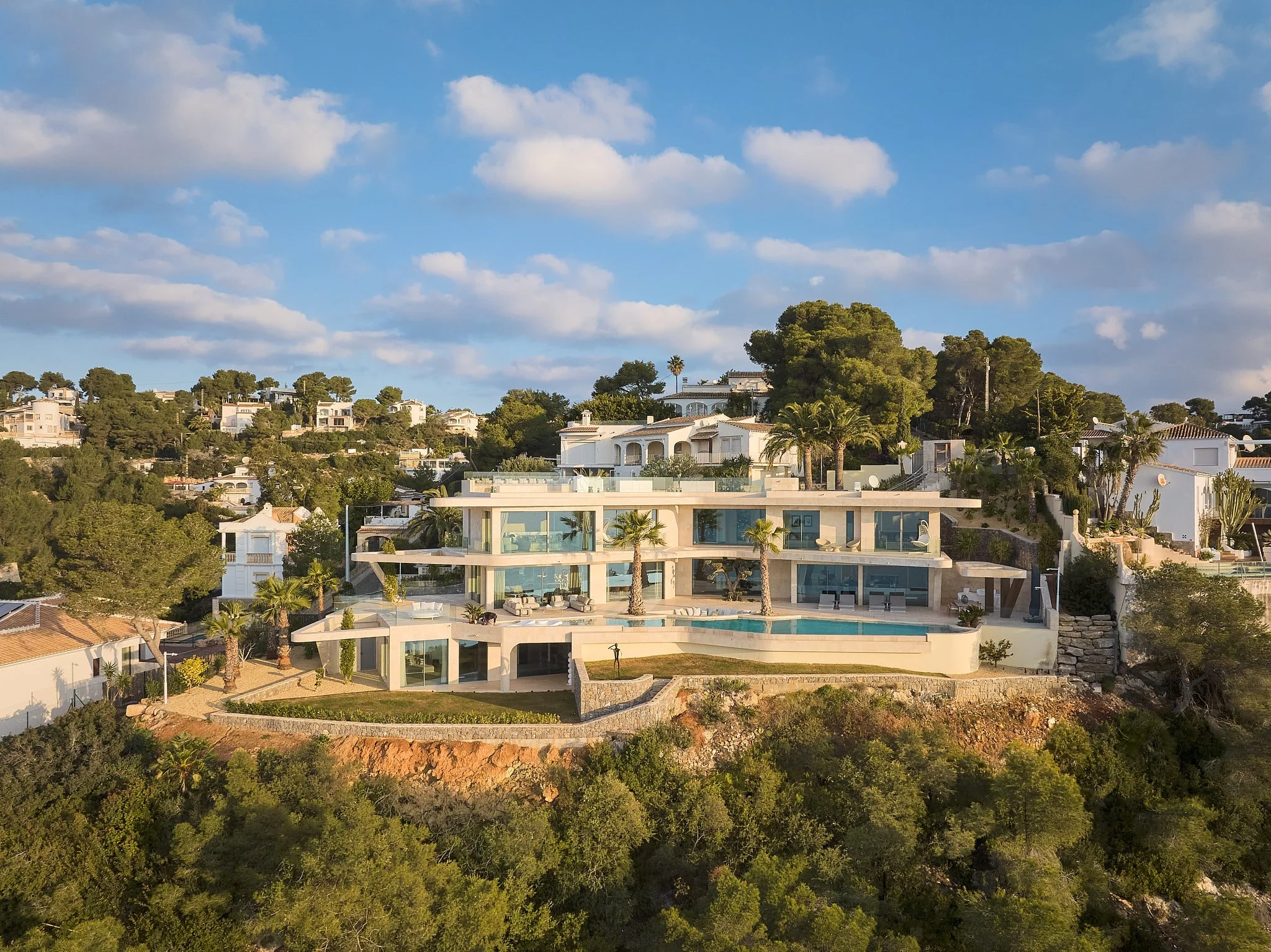 Casa moderna con varias terrazas y piscina en una colina, rodeada de árboles y casas blancas, con cielo despejado y algunas nubes.