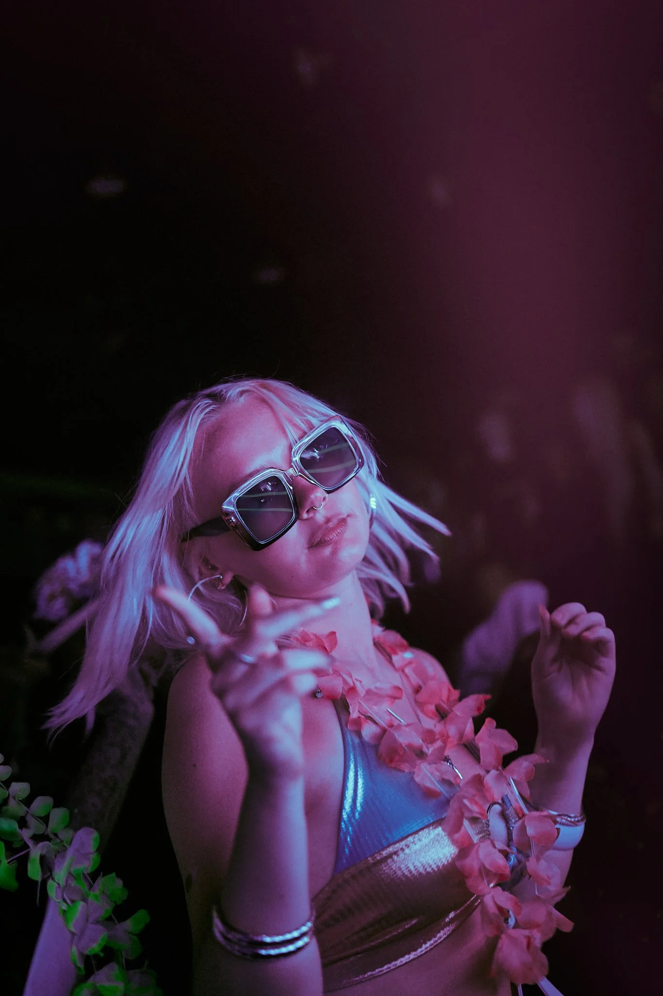 Joven mujer con gafas grandes plateadas, vestido dorado y collar de flores rosas, en un entorno con luces de discoteca y plantas verdes.