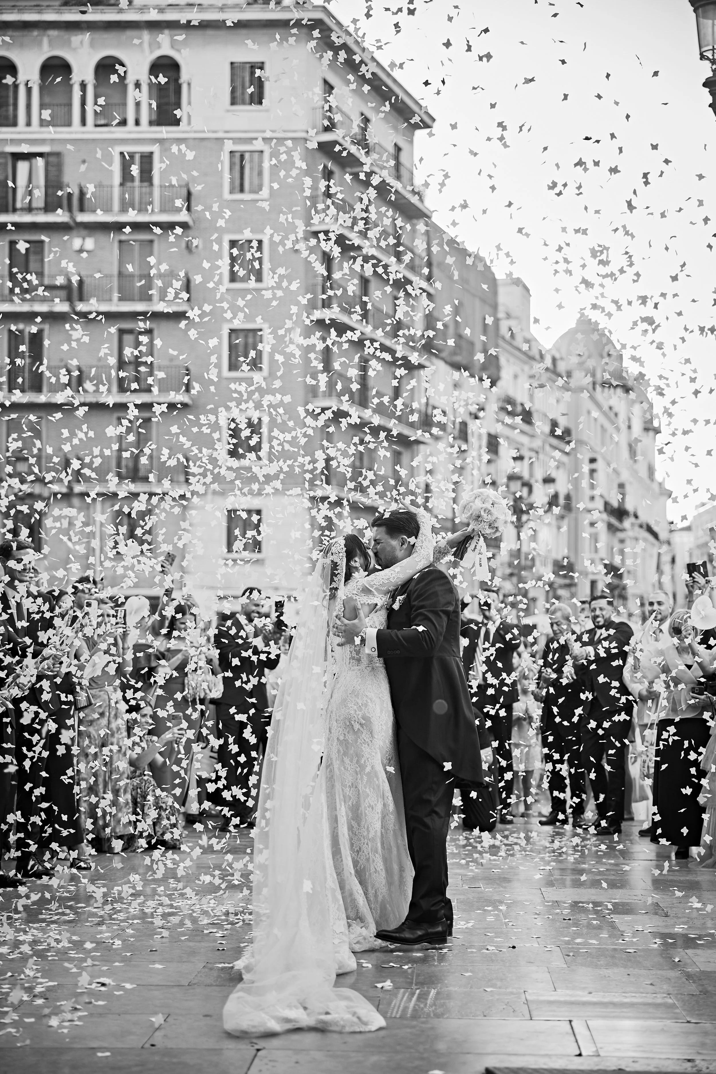 Pareja de novios bailando en una calle, rodeados de invitados, con confeti en el aire, en una celebración de boda al aire libre, en un entorno urbano con edificios de fondo, en blanco y negro.
