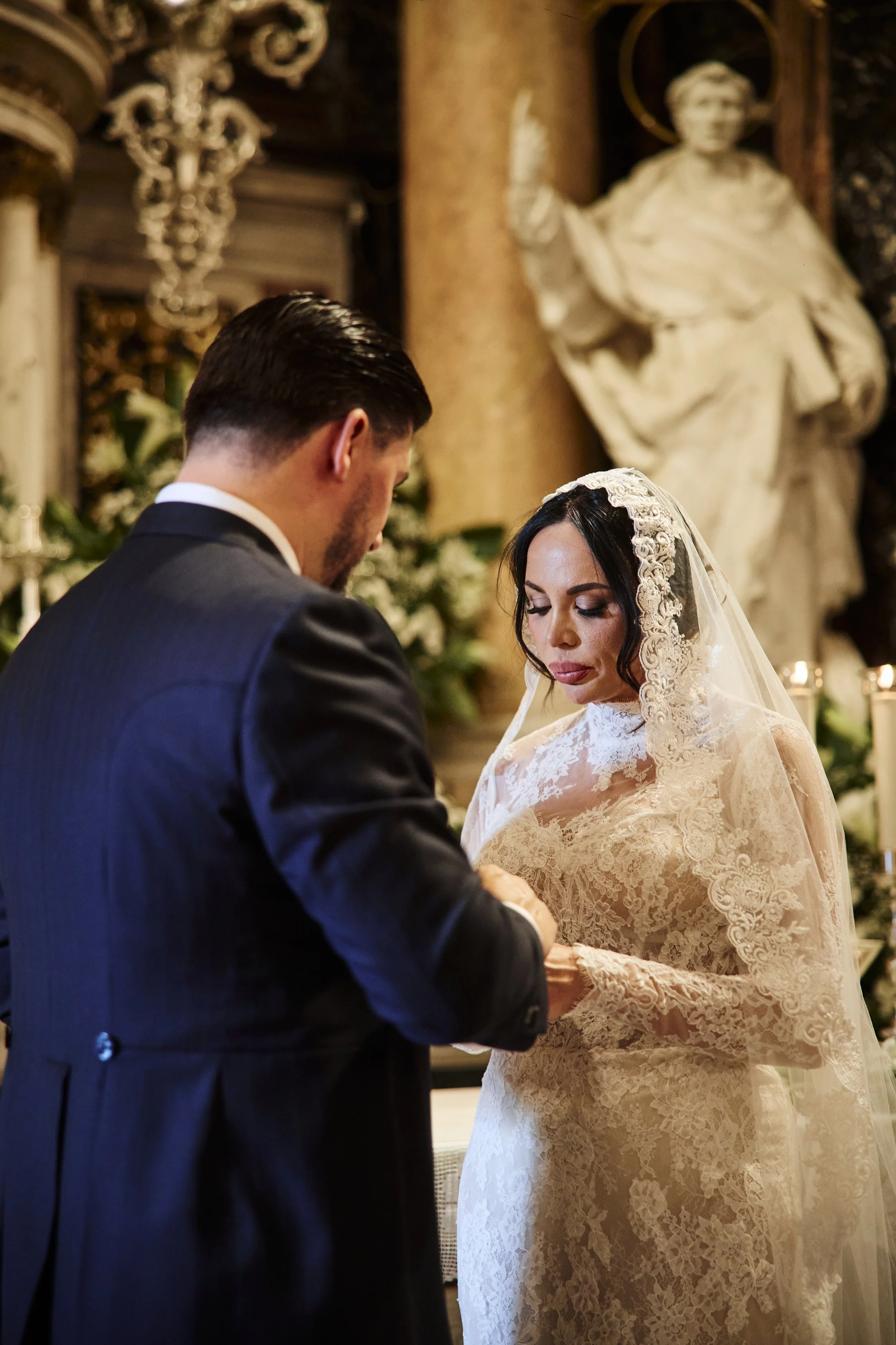 Una pareja en una ceremonia, con la mujer vestida de vestido de encaje y velo, y el hombre con traje oscuro, en un lugar decorado con columnas y una escultura de una figura humana en el fondo.