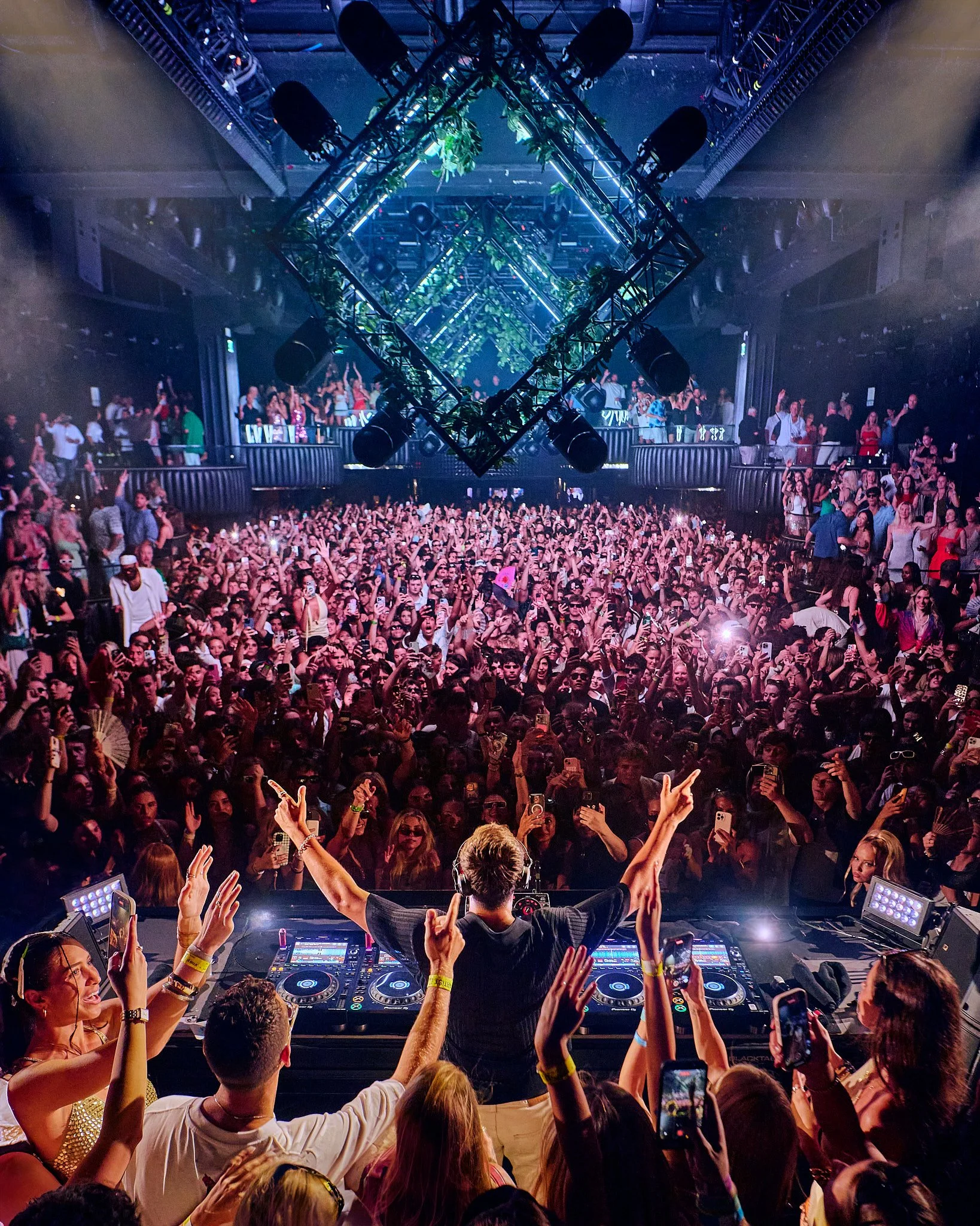 Una multitud en un club nocturno bailando y disfrutando de John Summit que toca en la cabina. La experiencia está llena de luces y energía.