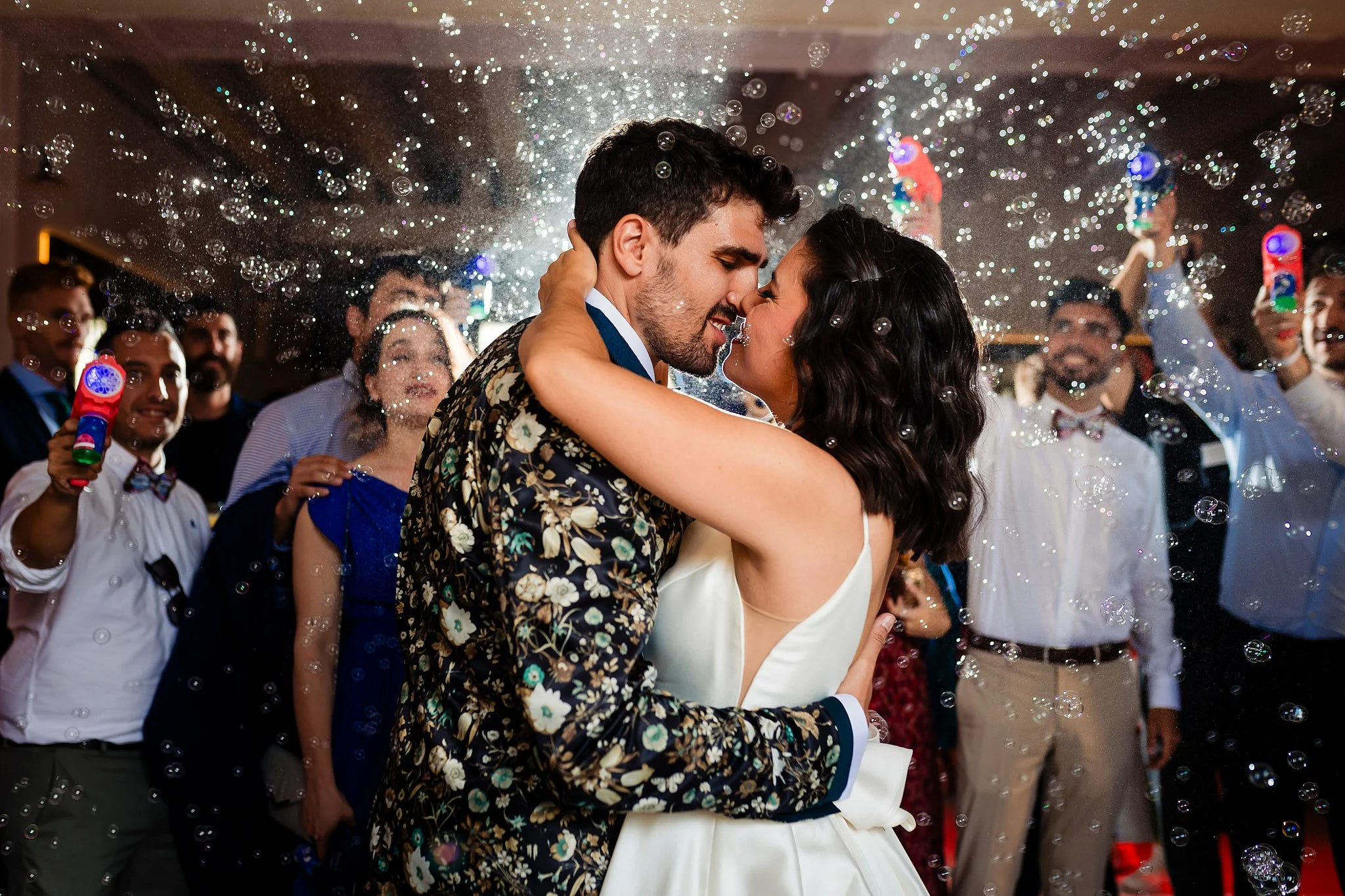 Pareja bailando en una boda con burbujas en el aire, rodeada de invitados que celebran y toman fotos.