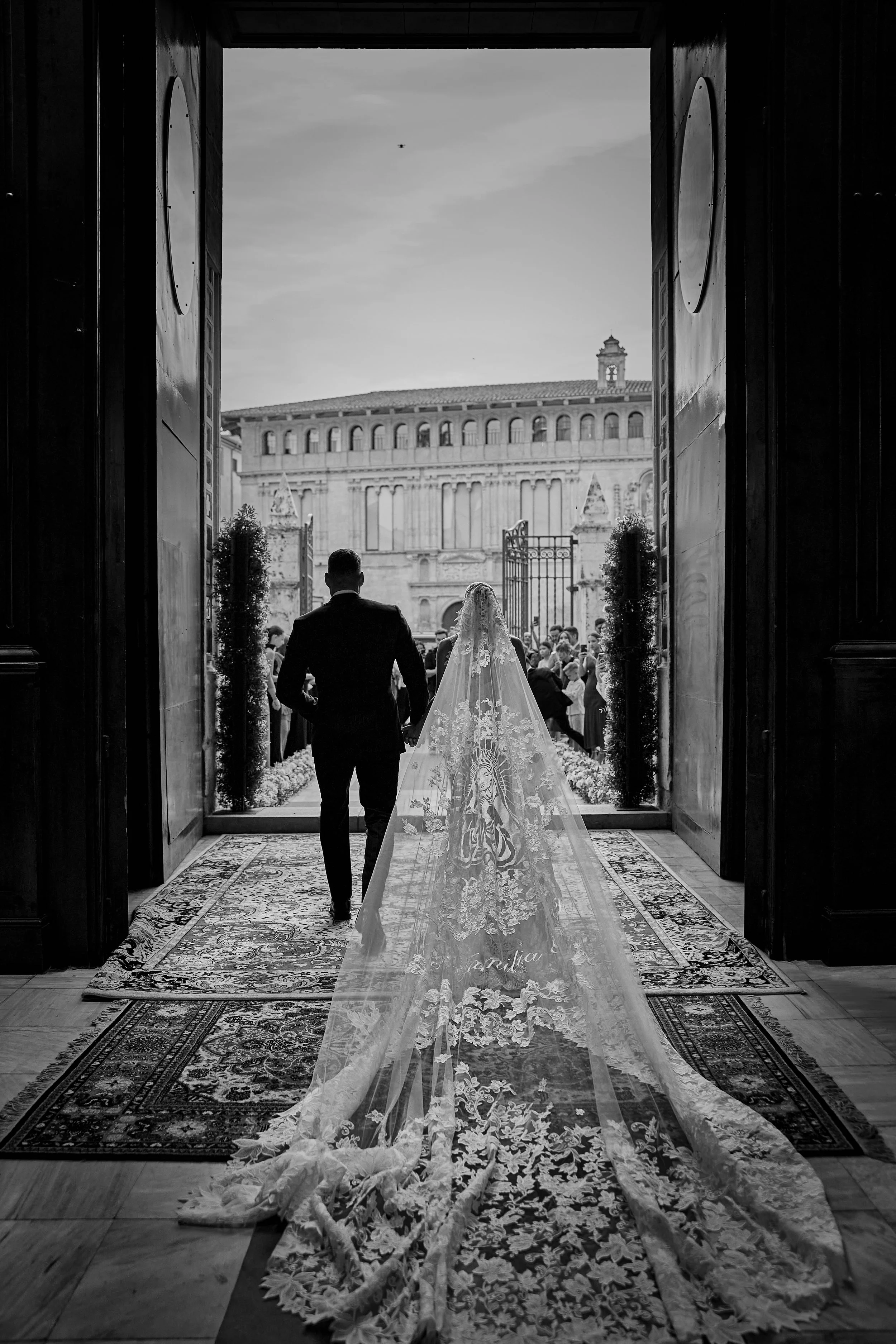 Una pareja de recién casados entrando a una iglesia para su boda, vista desde la entrada, con una multitud de invitados afuera y un edificio antiguo en el fondo; en blanco y negro.
