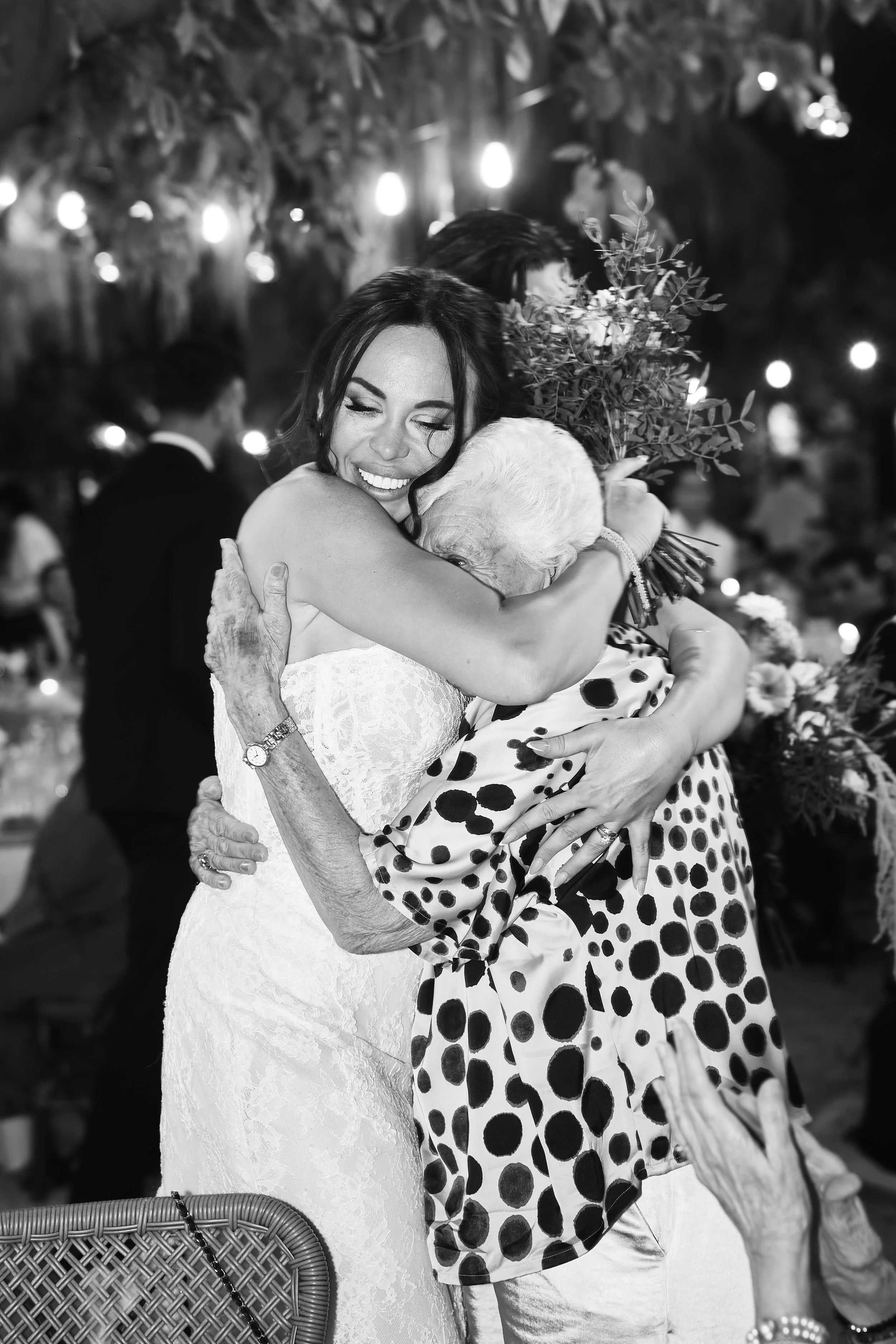 Mujer con vestido blanco abrazando a una anciana que lleva un vestido con lunares y sostiene un ramo de plantas, en un evento social con luces decorativas en el fondo.