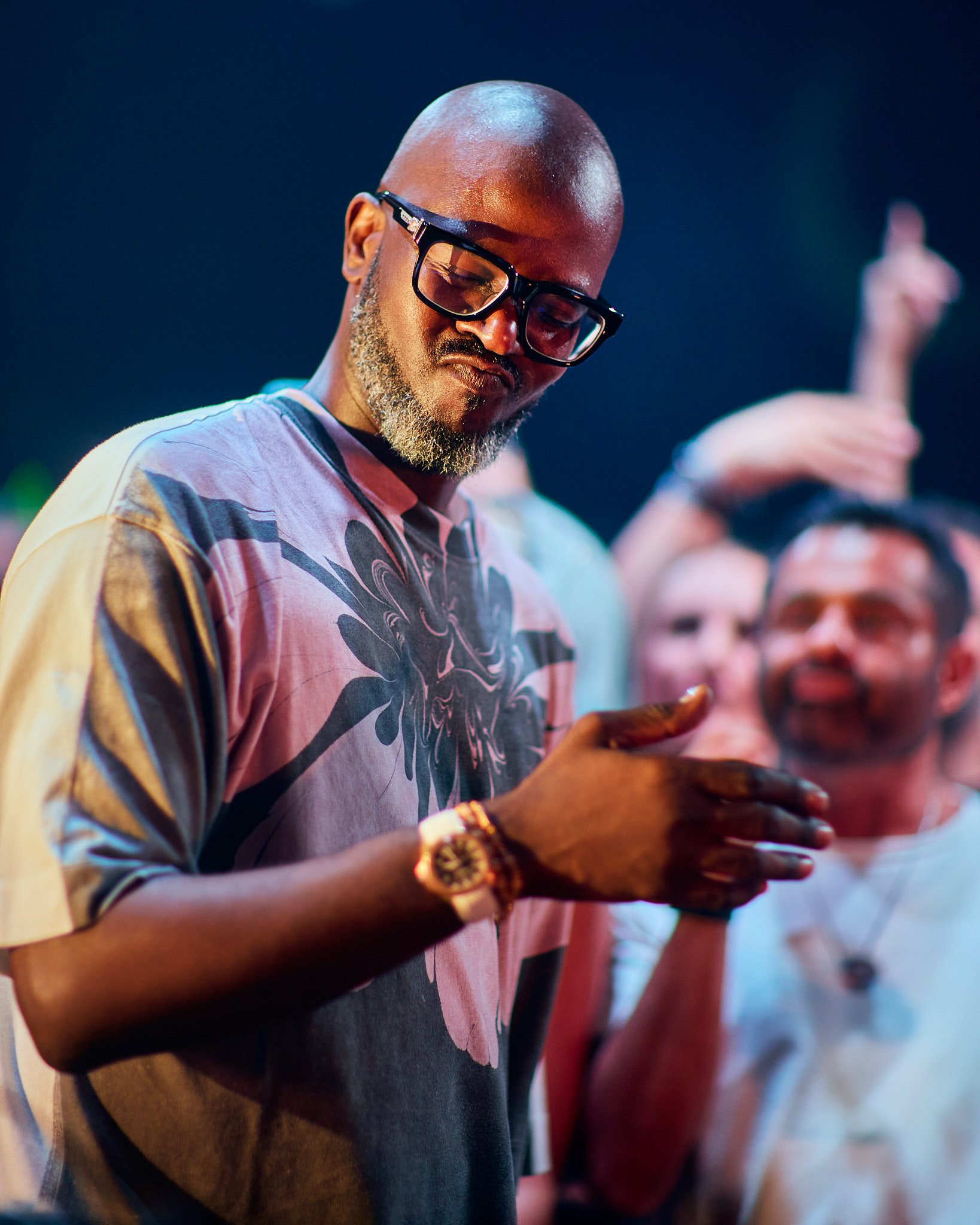 Hombre con gafas y barba en un evento, usando una camiseta con diseño colorido, mirando su teléfono móvil con varias personas en el fondo.