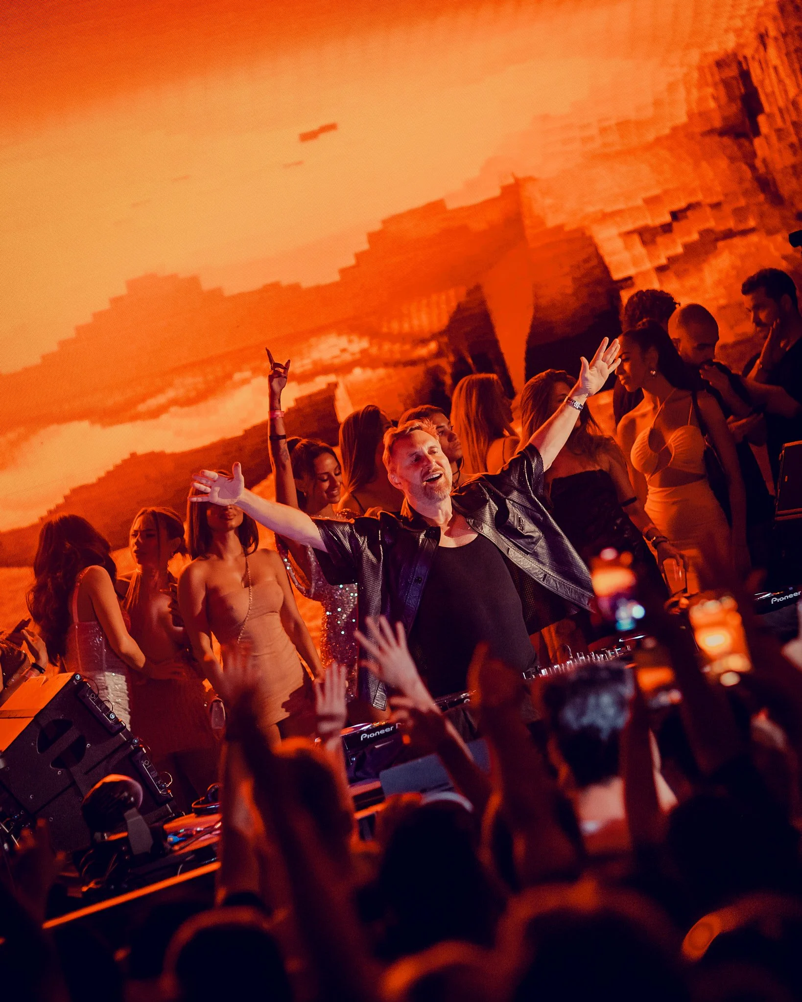 David Guetta con chaqueta de cuero en un escenario con muchas personas bailando y disfrutan, con fondo de pantallas con imágenes en tonos anaranjados y rojos.