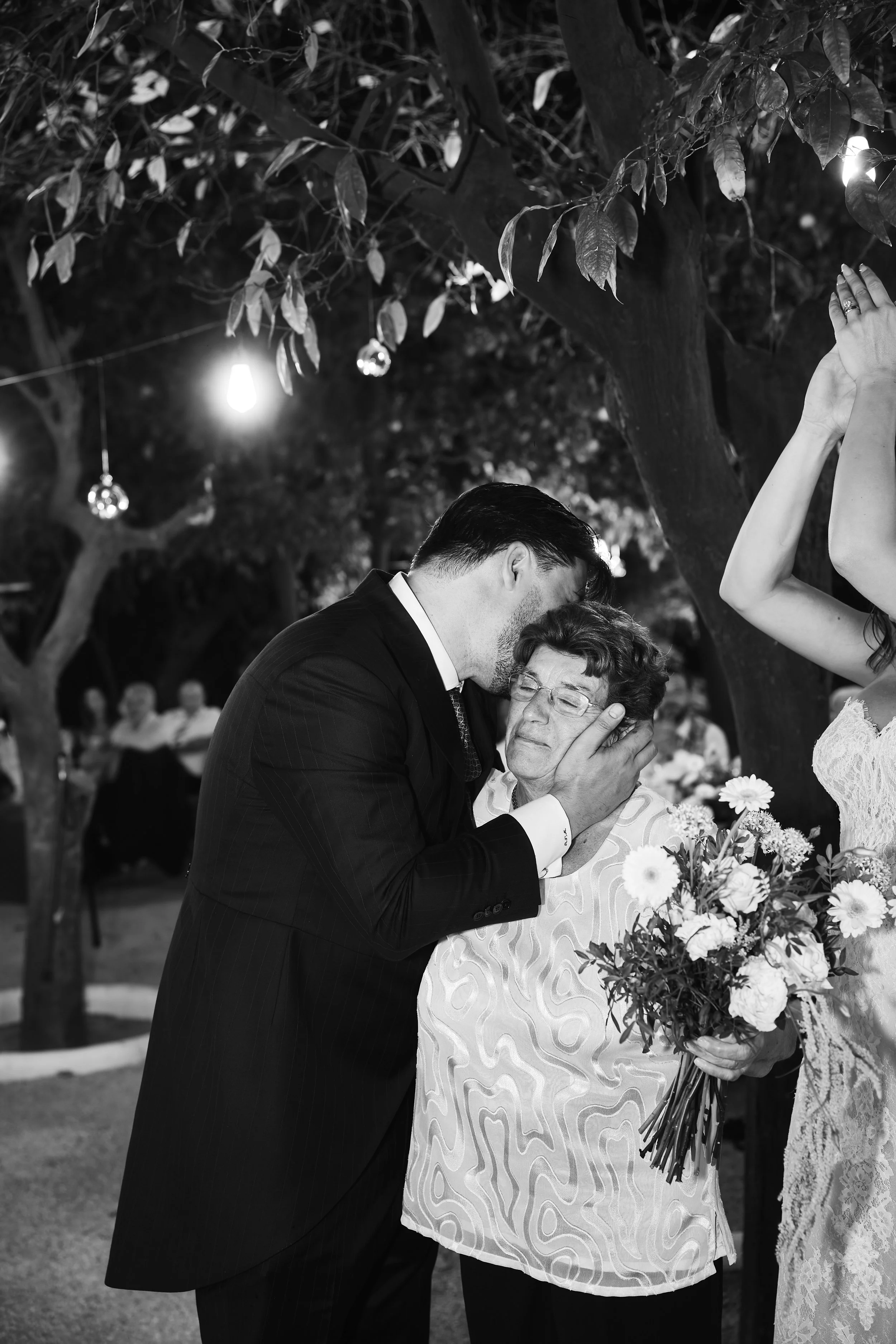 Un hombre y una mujer mayores se abrazan y comparten un momento emotivo con un fondo de árboles y luces de ambiente en una celebración al aire libre.